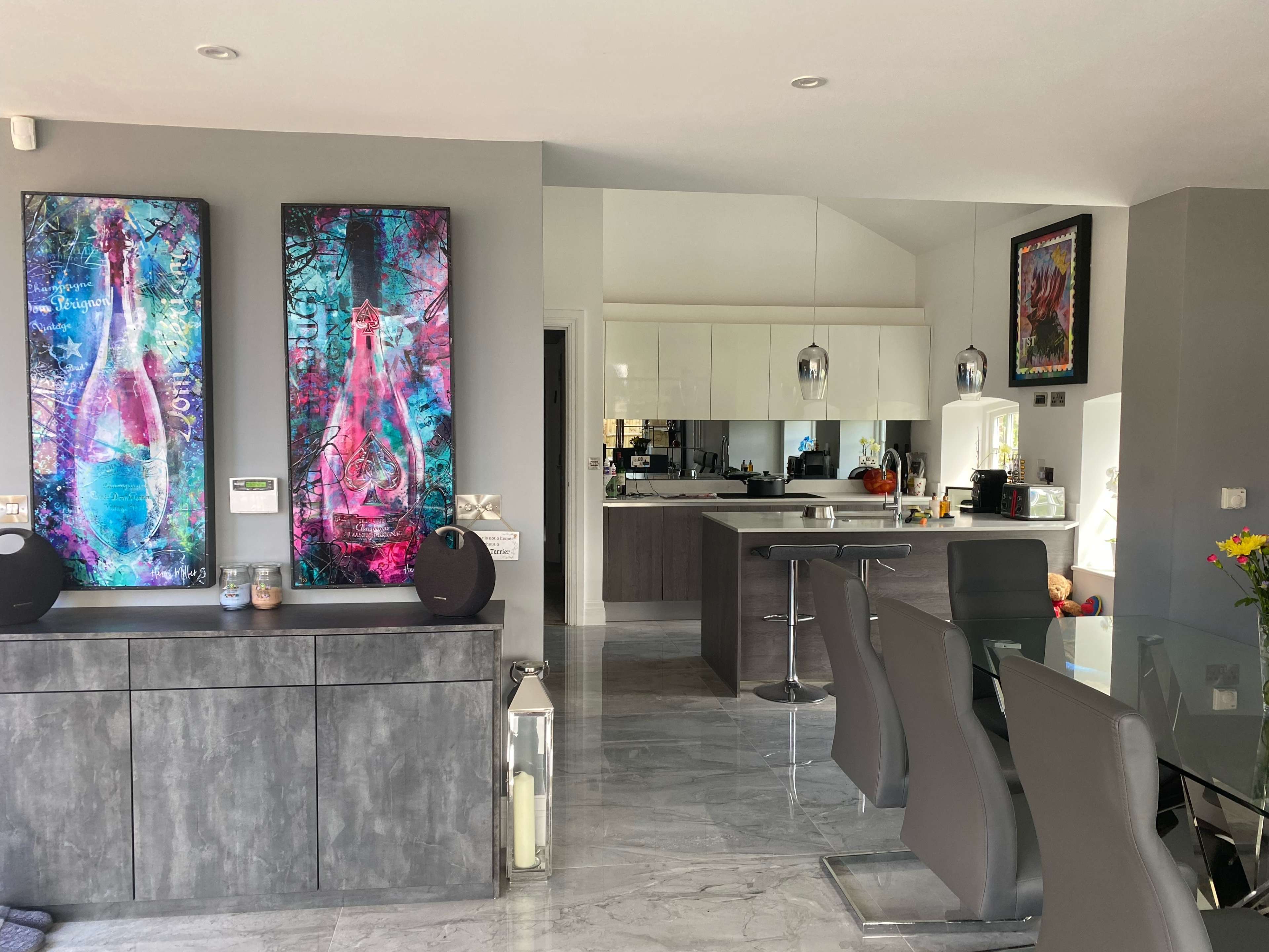 A modern kitchen and dining area featuring gray cabinetry, abstract artwork on the walls, and a glass dining table with gray chairs.
