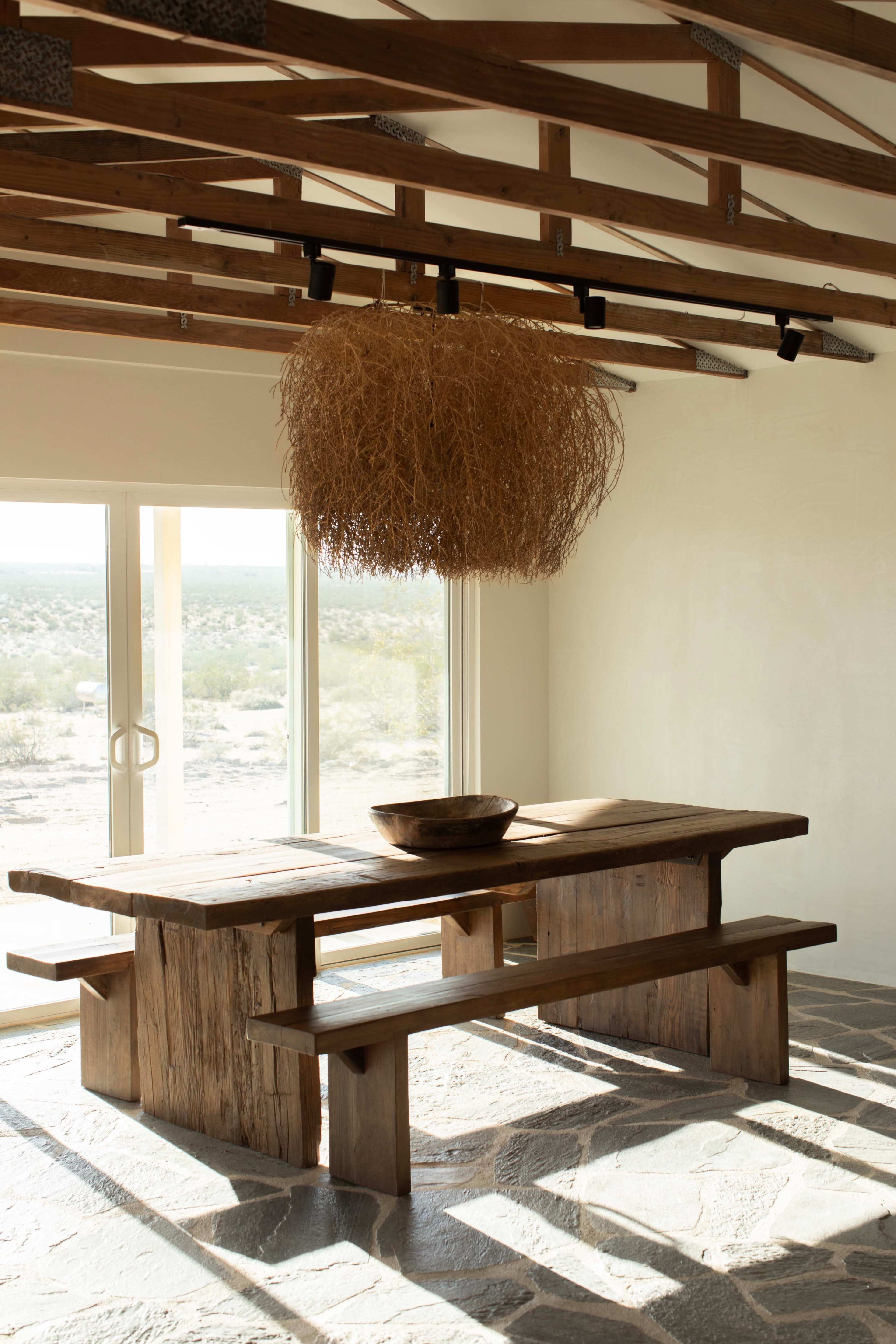 A wooden dining table with matching benches is centered in a bright room featuring a large light fixture and a view of a desert landscape through sliding glass doors.