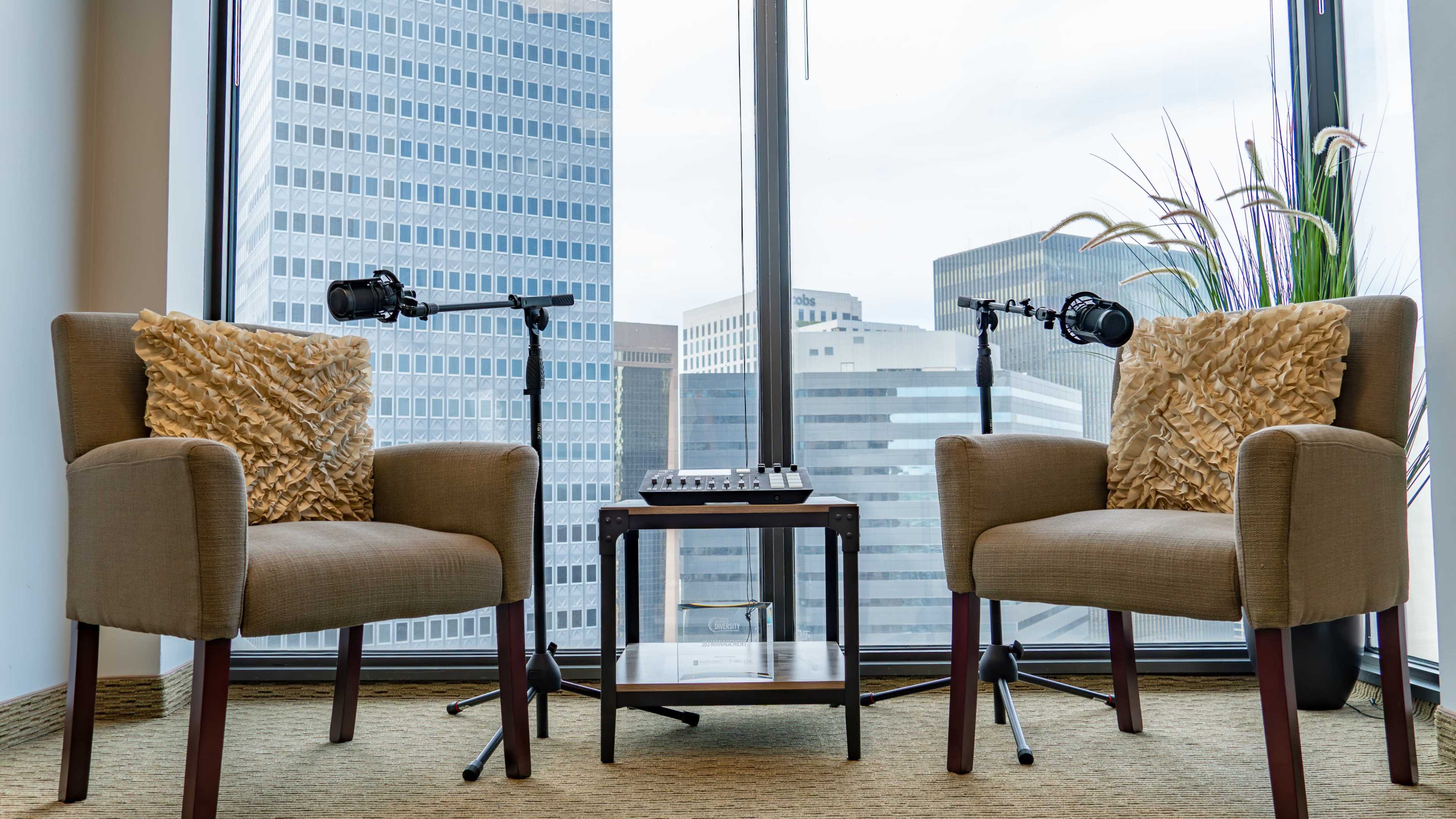 Two upholstered chairs with decorative pillows are positioned near a window, with a small table and microphone stands between them, overlooking a city skyline.