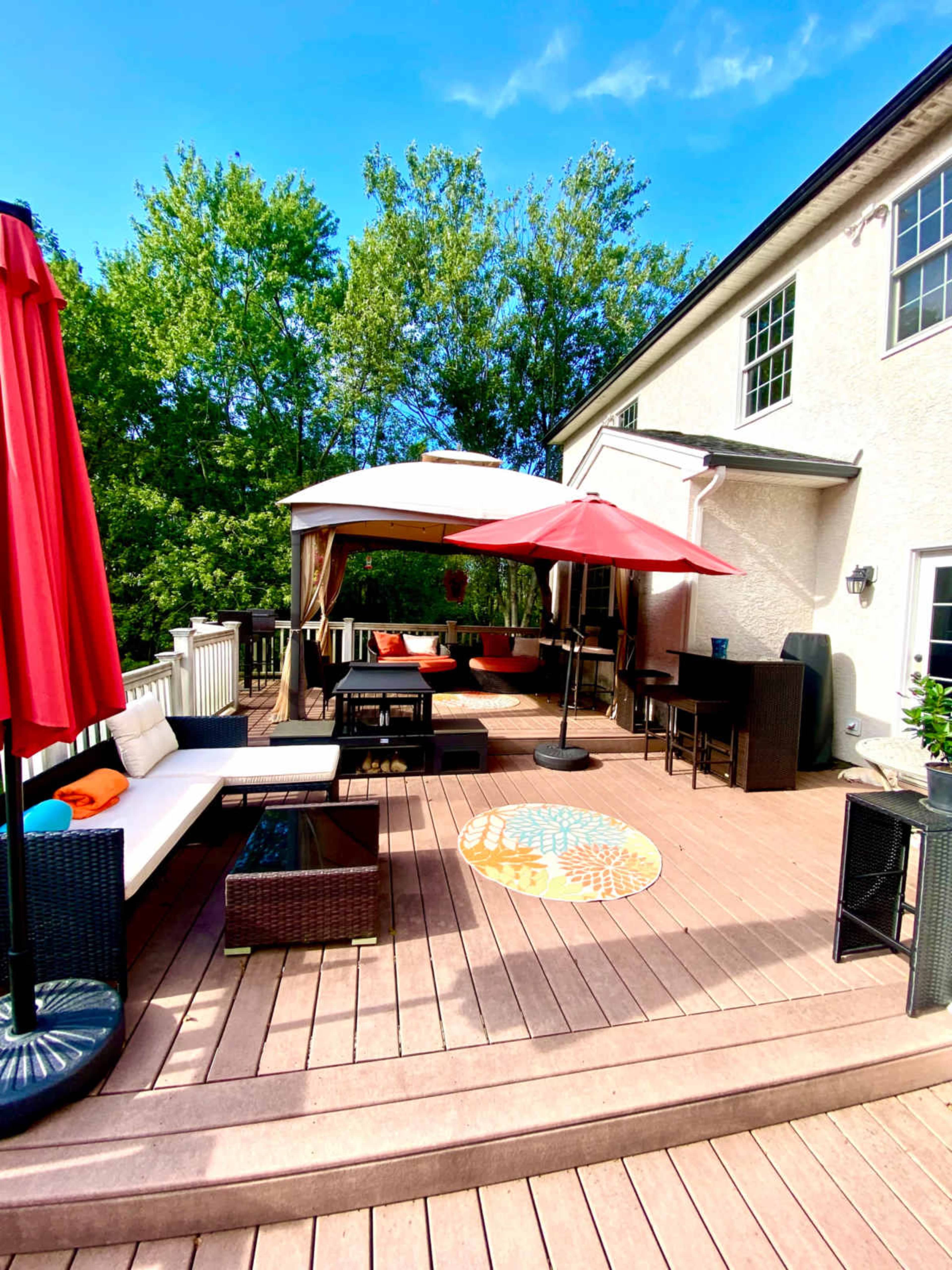 The image shows a wooden deck area outfitted with outdoor seating, umbrellas, and a canopy, surrounded by trees.
