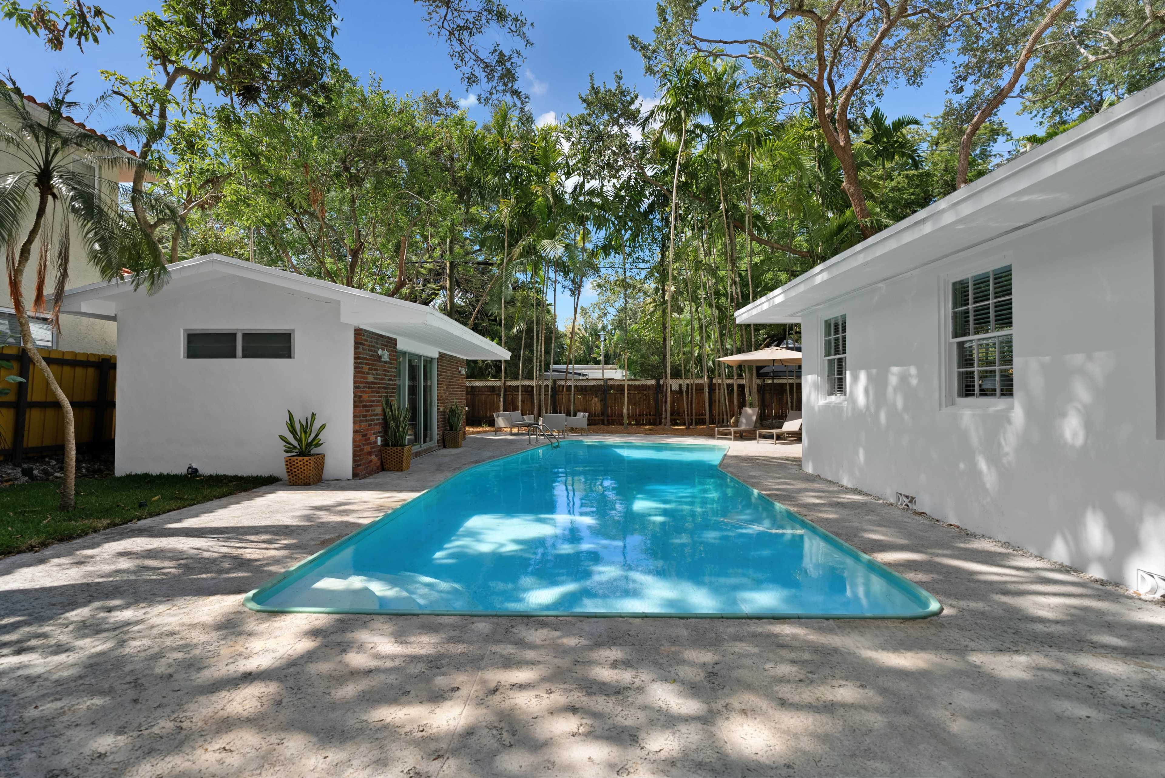 The image shows a brightly lit outdoor area featuring a swimming pool surrounded by white buildings and lush greenery.