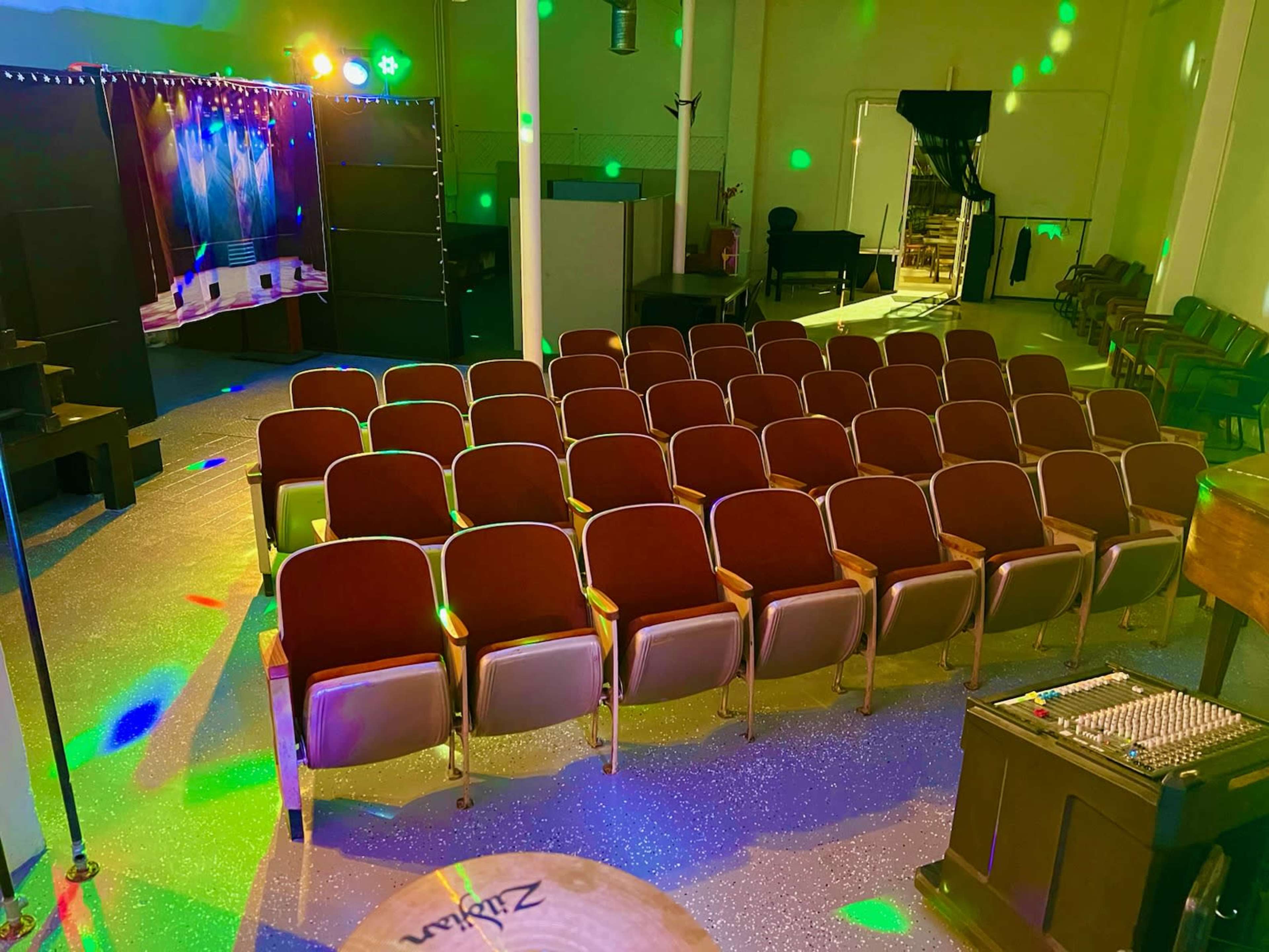 An empty auditorium with rows of maroon seats arranged in front of a stage, illuminated by colorful lights.