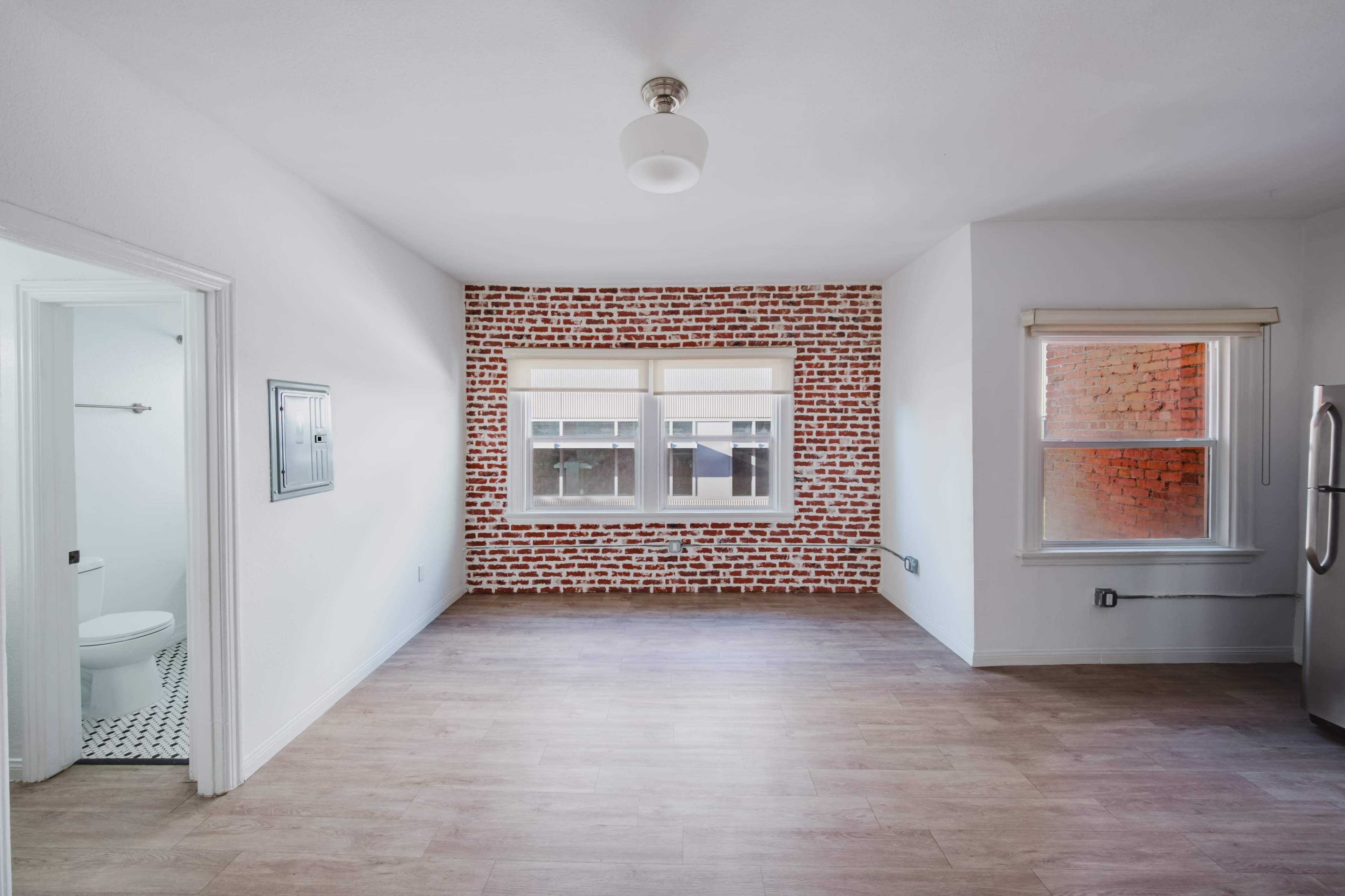 The image shows a spacious room with a brick accent wall, large windows, and a view of a bathroom door in the corner.
