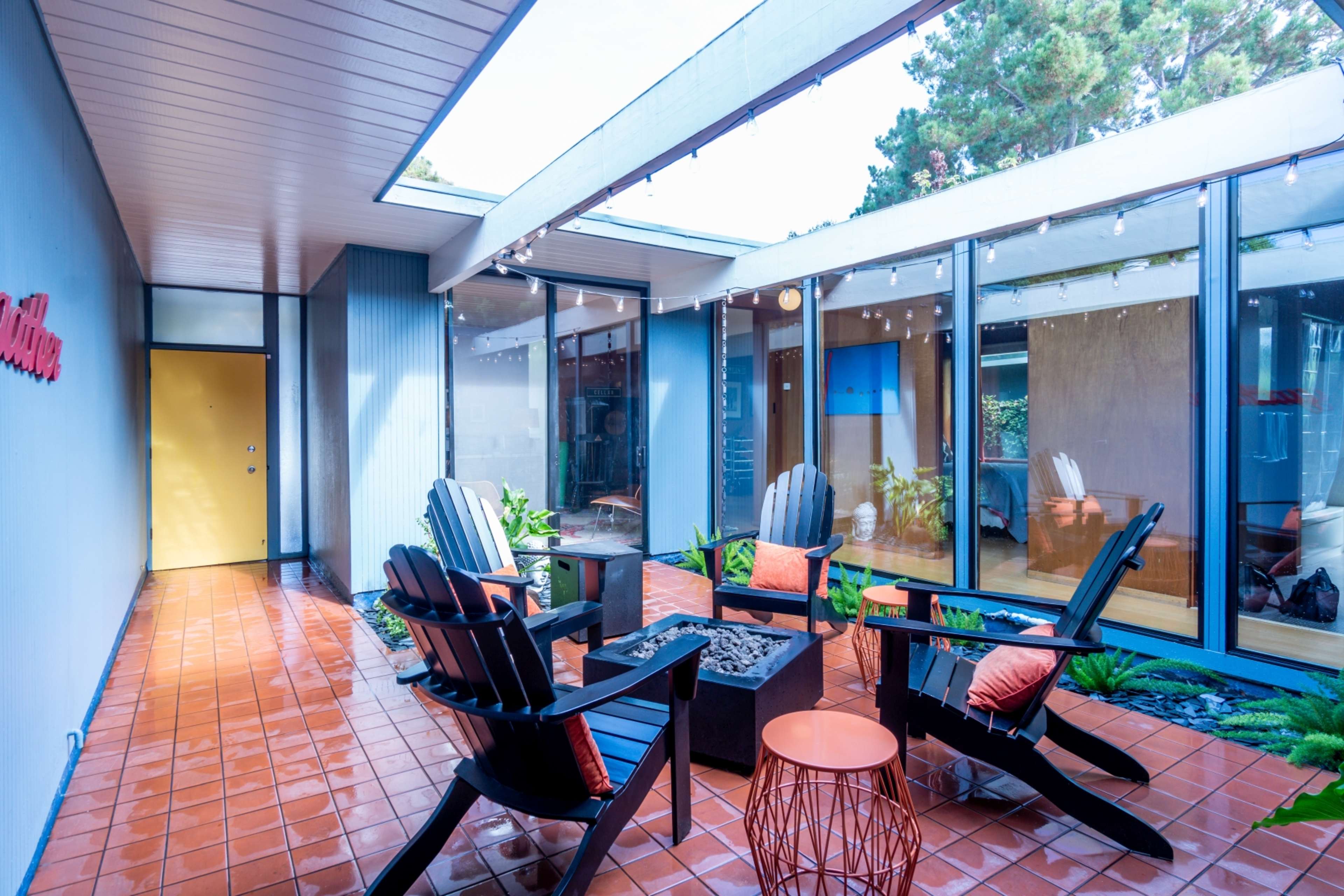 The image shows a modern courtyard with four black Adirondack chairs arranged around a fire pit, surrounded by greenery and large windows.