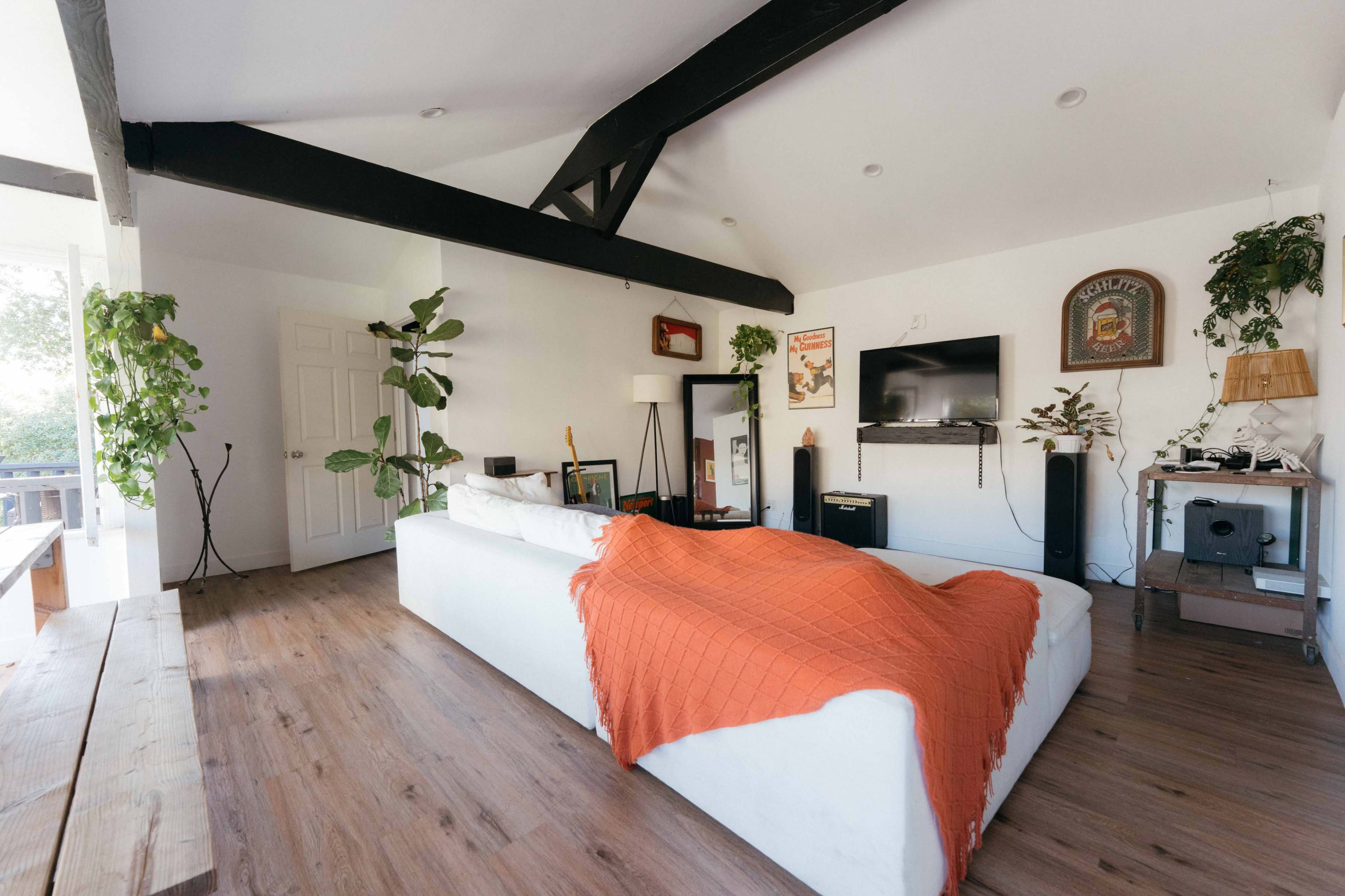 A modern living room features a white sectional sofa adorned with an orange throw blanket, surrounded by indoor plants and wooden accents, with a television mounted on the wall.