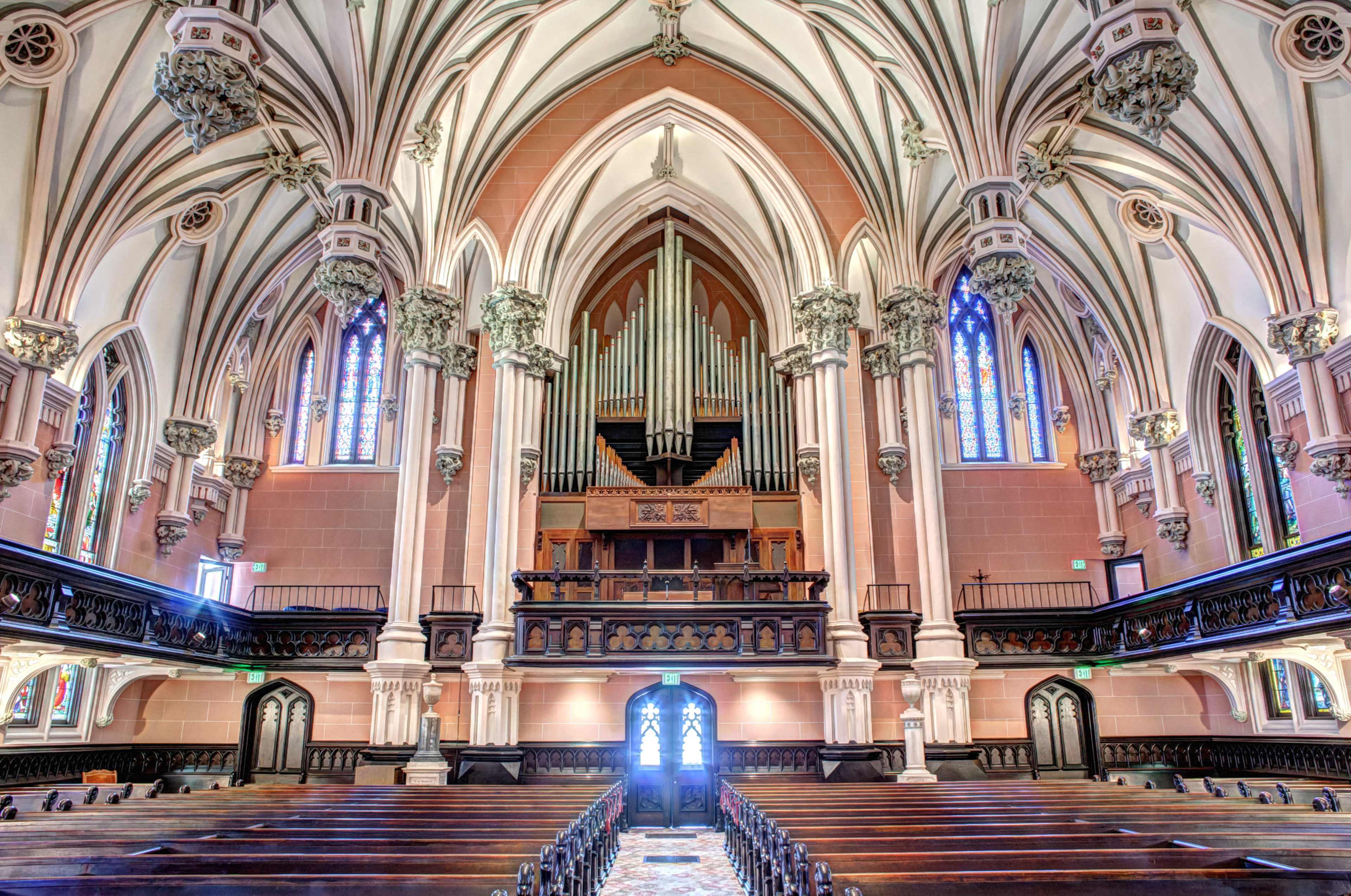 The interior of a grand church features tall, arched ceilings, ornate columns, an organ at the back, and rows of wooden pews facing a central entryway.
