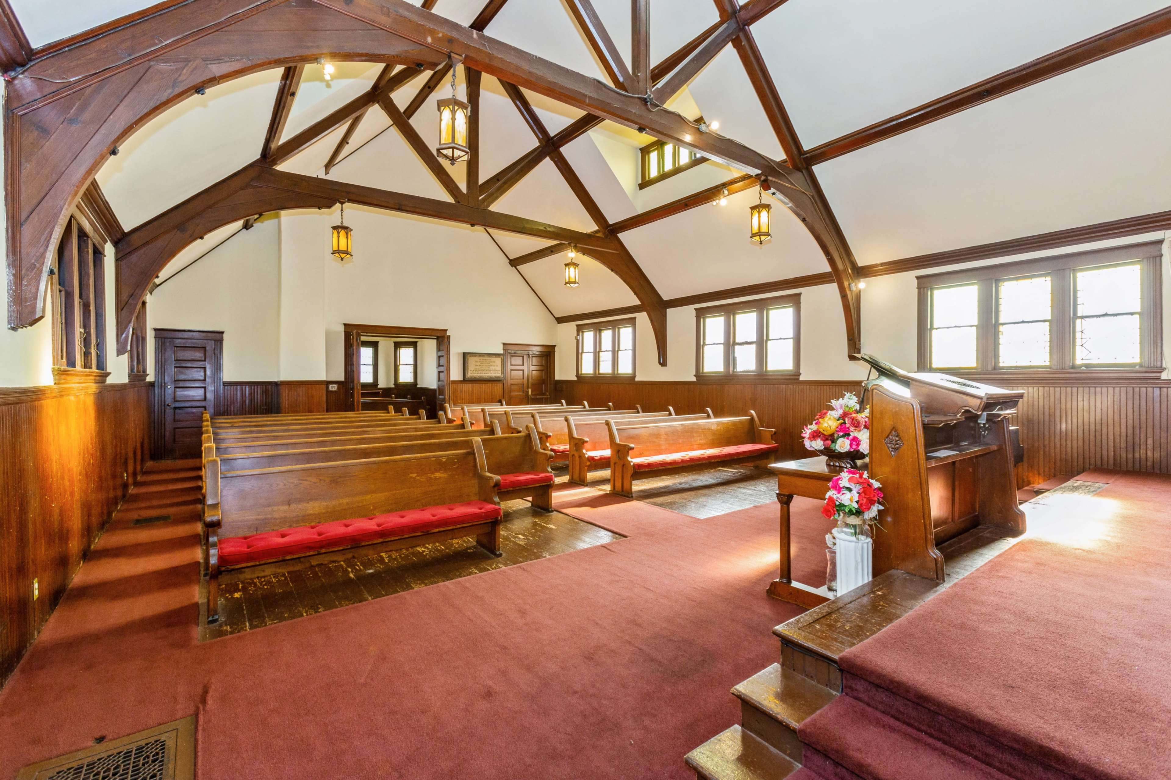 The interior of a church features wooden beams, rows of wooden pews with red cushions, and a floral arrangement near the pulpit.