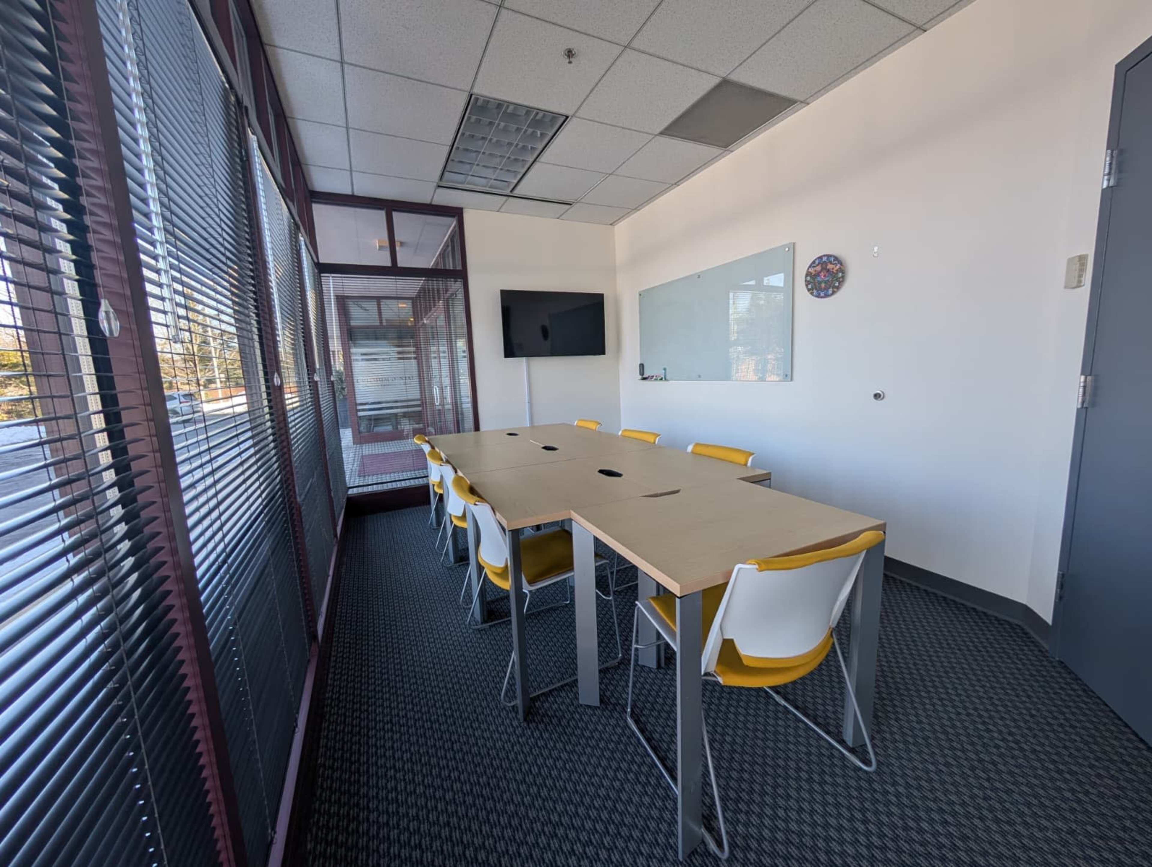 The image shows a modern conference room with a long table surrounded by yellow and white chairs, a wall-mounted screen, and large windows covered with blinds.