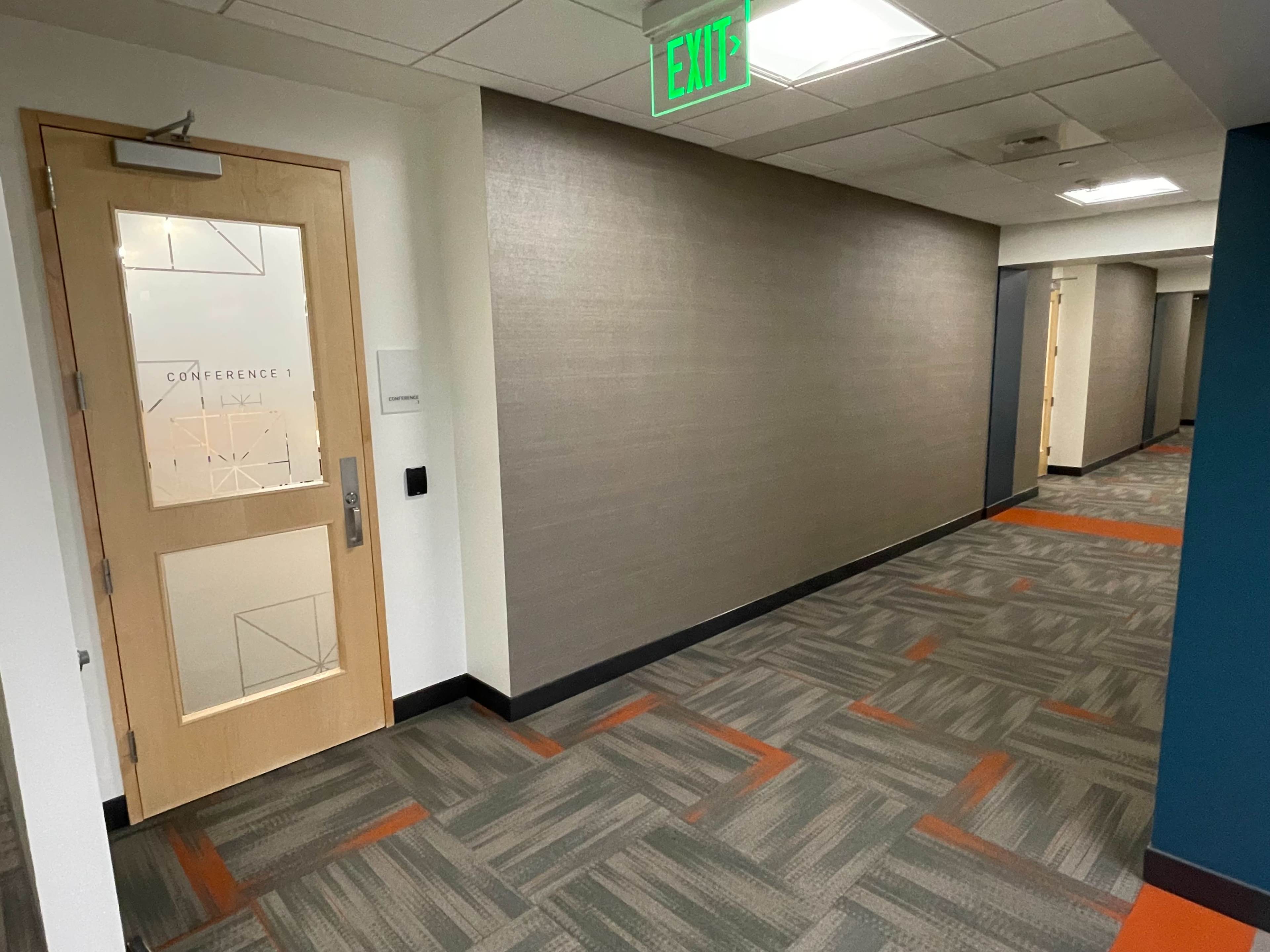 A corridor with a wooden door labeled "CONFERENCE 1" and a green exit sign above, lined with light gray walls and patterned carpet.