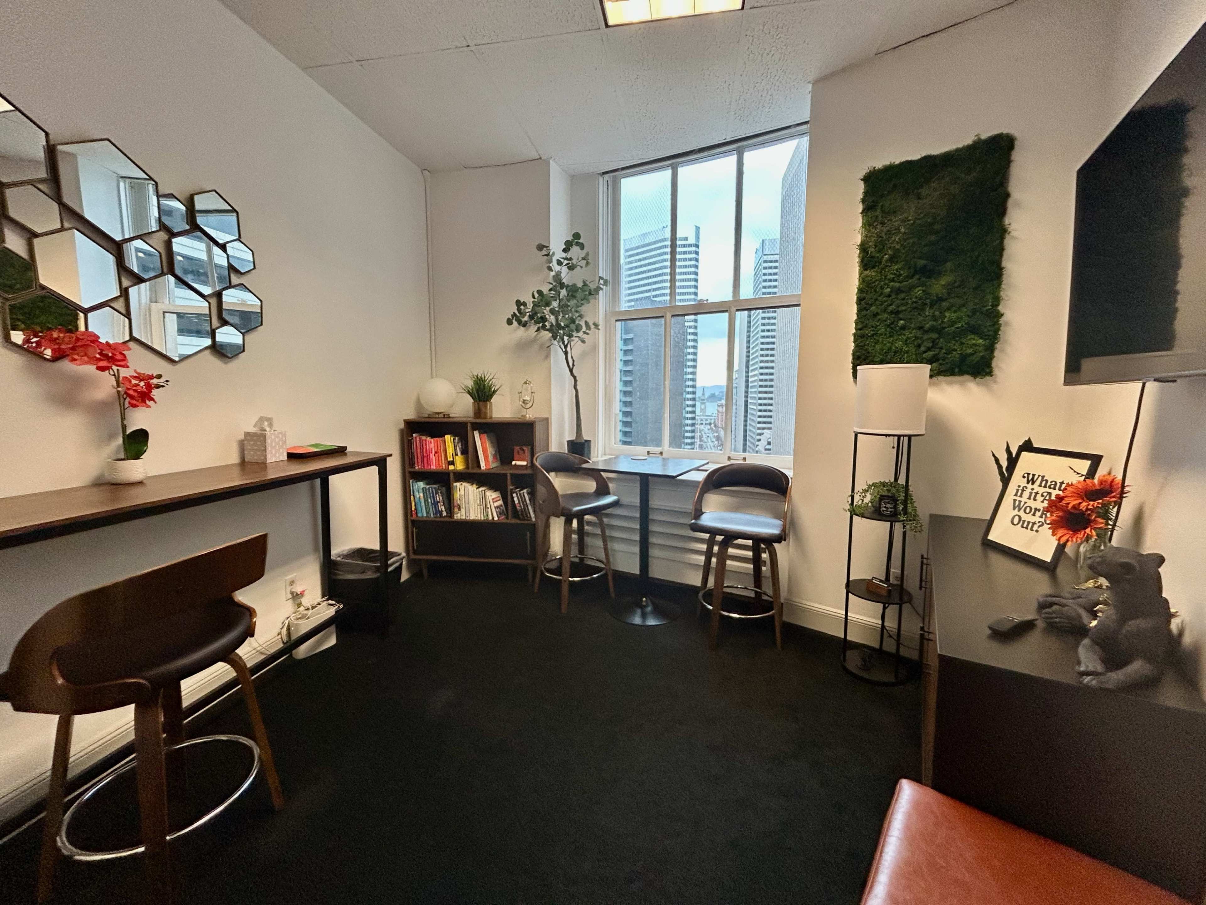 The image shows a small office space with a window overlooking tall buildings, featuring shelving with books, plants, and a cozy seating area.