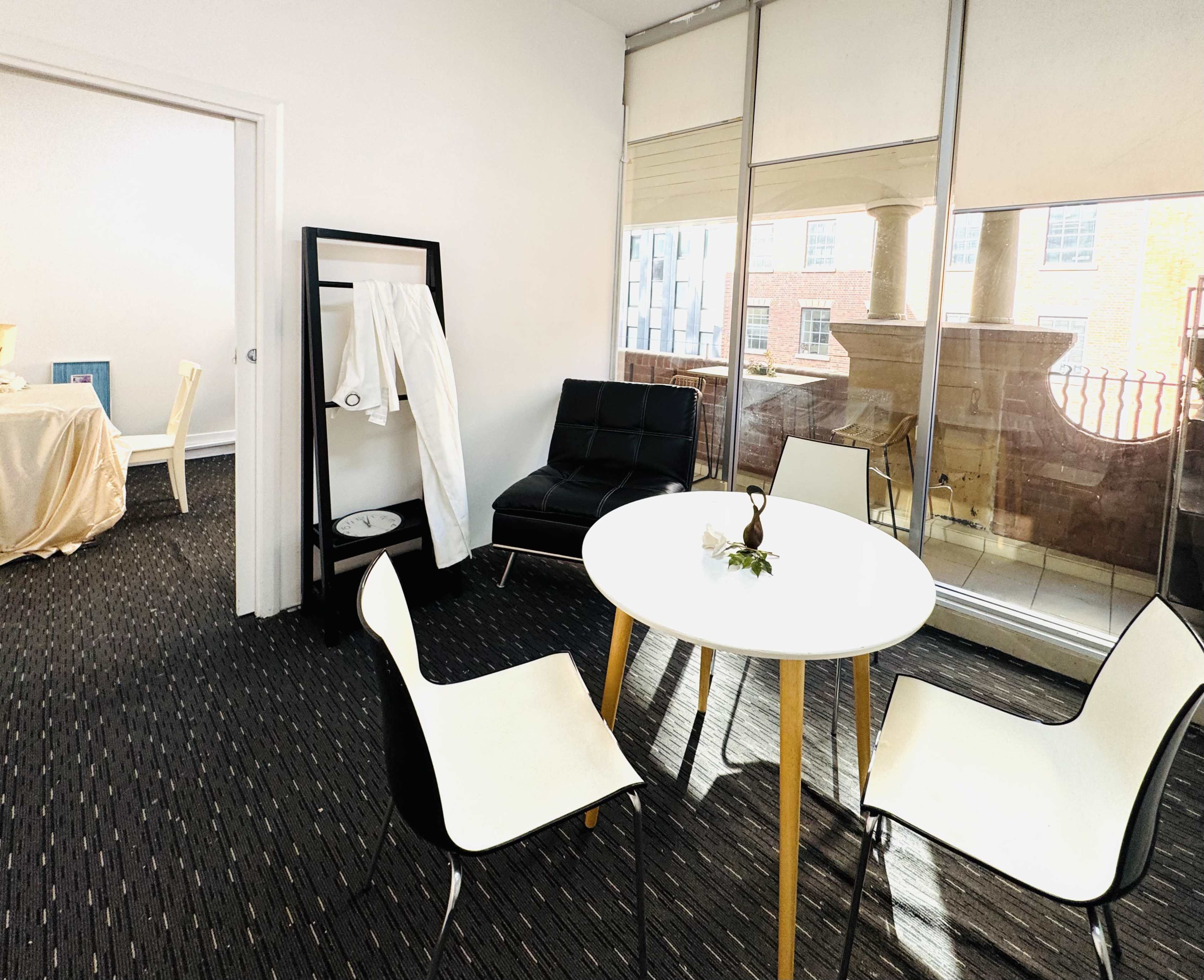 The image depicts a modern interior space featuring a round table with four chairs, a black seating area, and a clothing rack, with large windows offering a view outside.