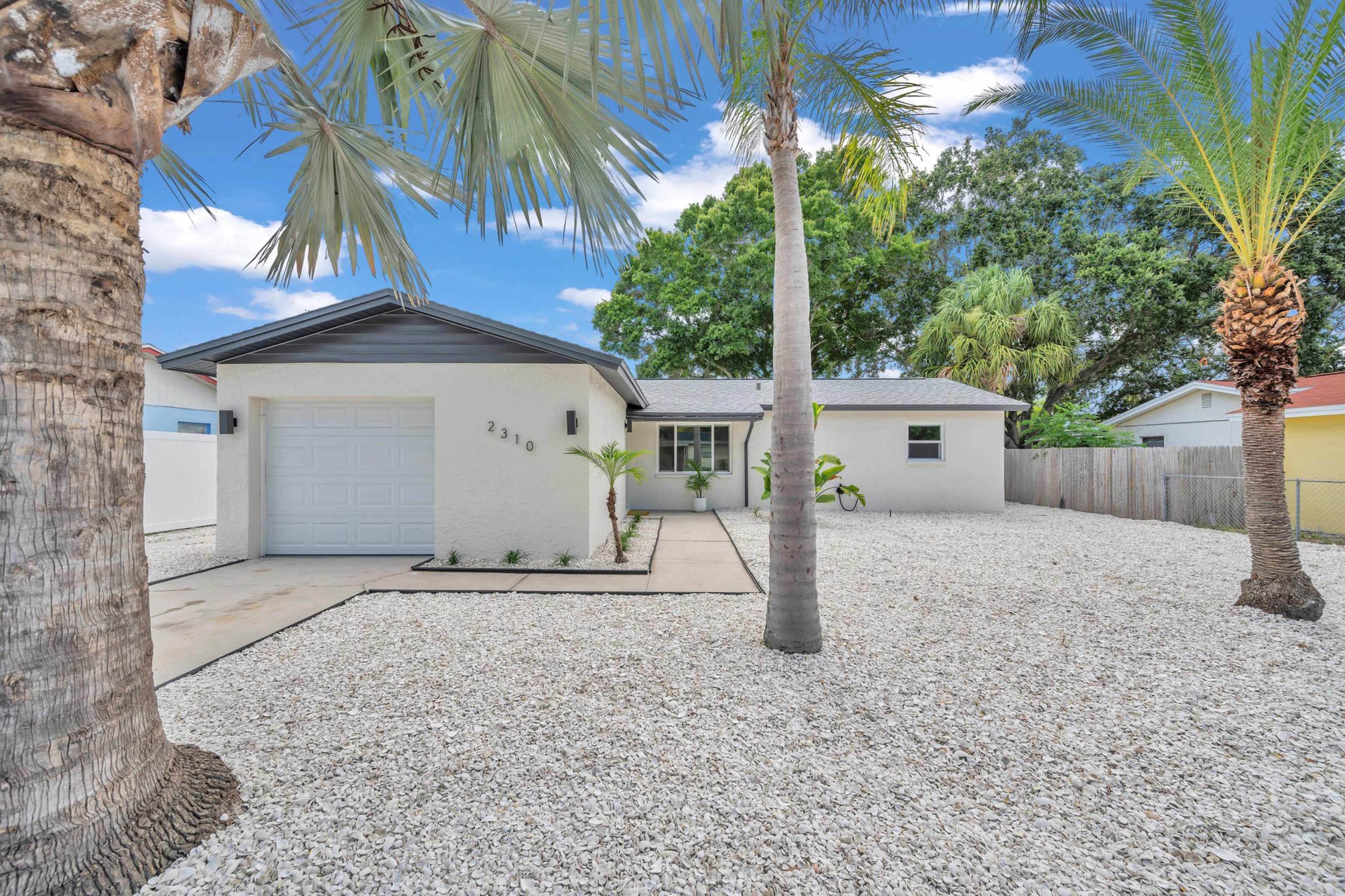 A single-story house with a garage, surrounded by a gravel yard and palm trees.