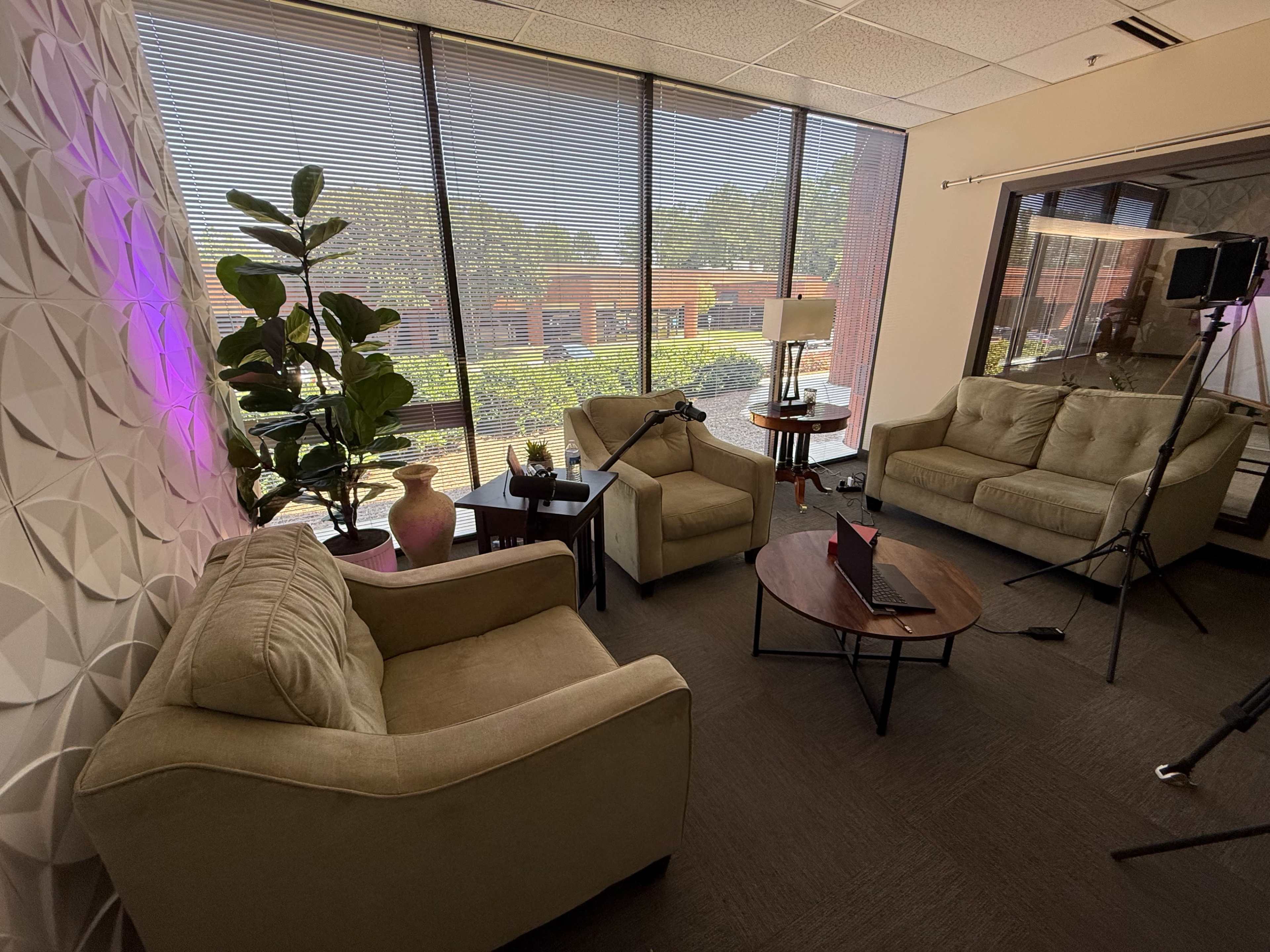 The image shows a cozy seating area with two sofas, a small table, and a plant, illuminated by natural light from large windows.