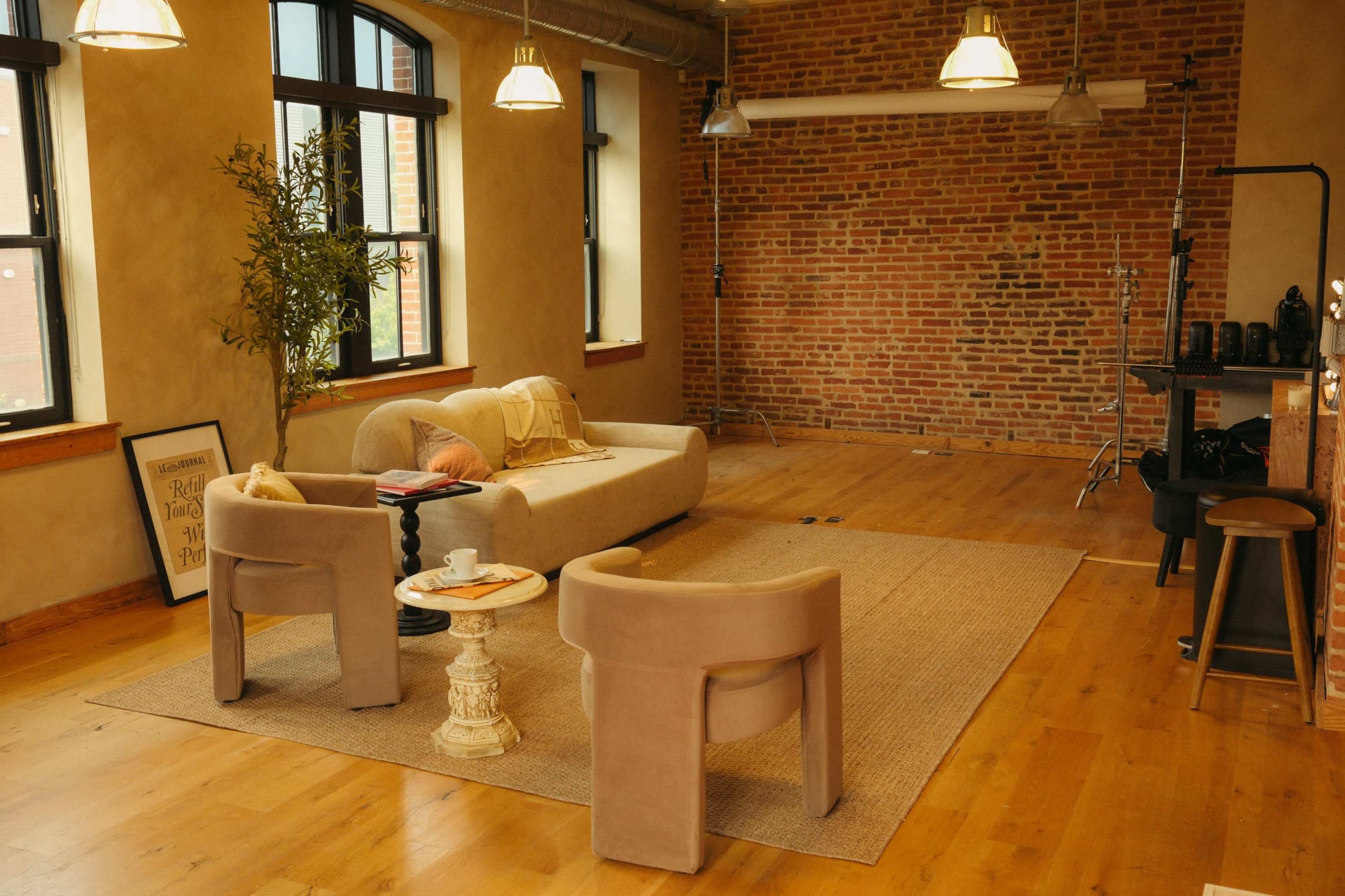 The image shows a spacious, well-lit room with a brick wall, a light-colored sofa, two armchairs, a small table, and wooden flooring.