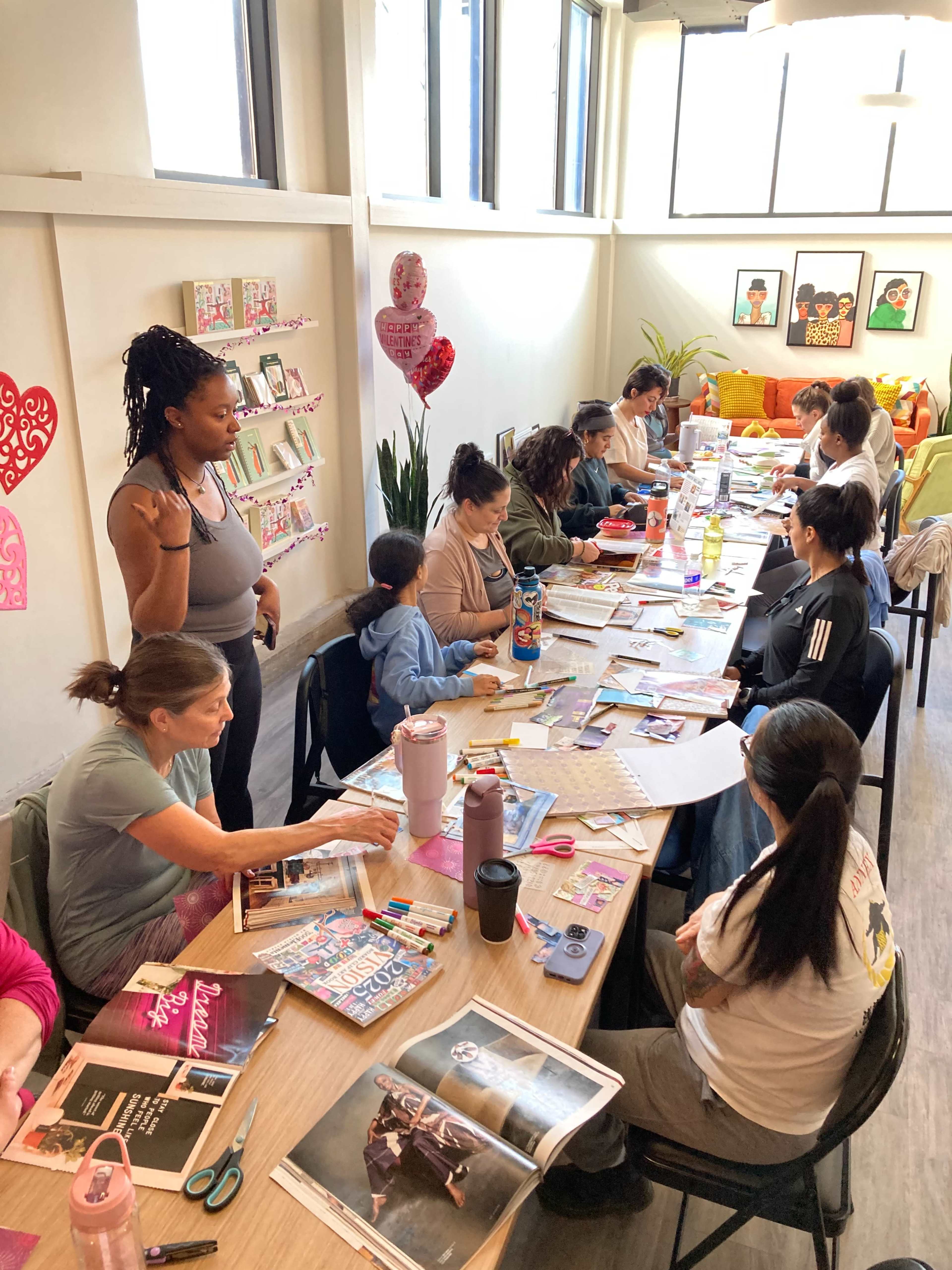 A group of people are engaged in a crafting session at a long table, creating vision boards using magazines, scissors, and various art supplies in a brightly lit room.