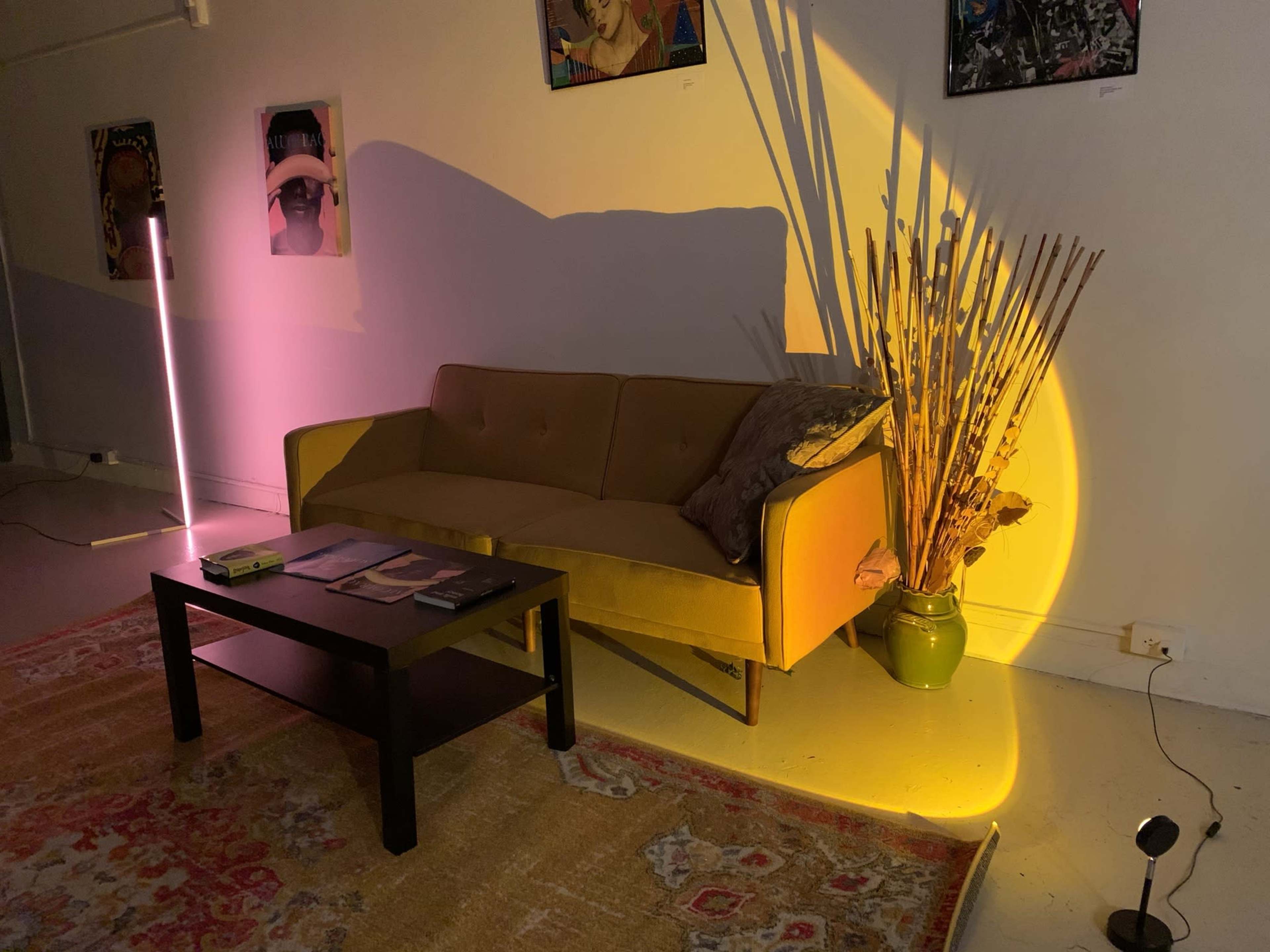 A yellow sofa positioned next to a table with magazines, surrounded by a colorful rug and a tall, decorative plant illuminated by soft lighting.