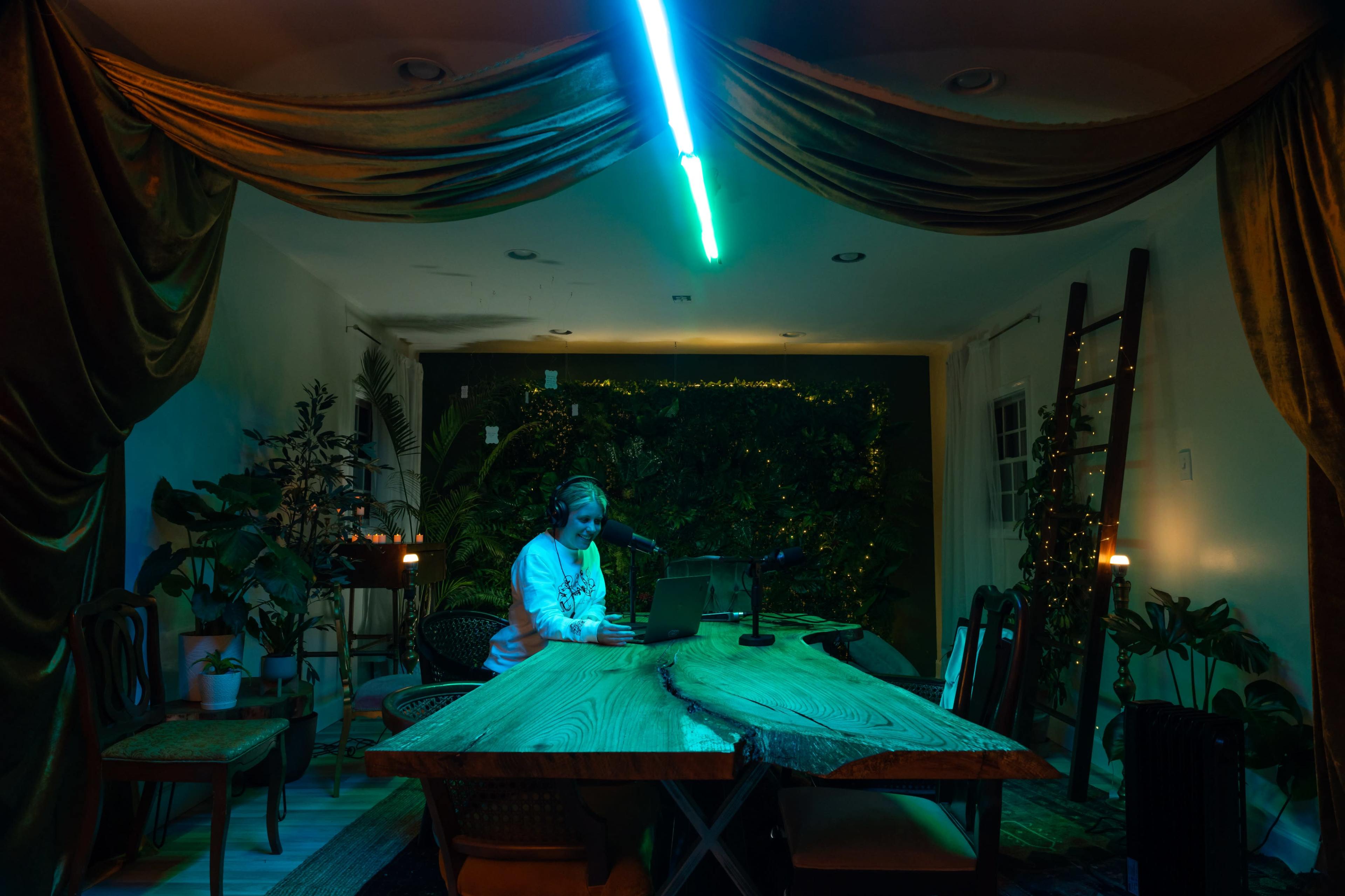 A person sits at a wooden table in a dimly lit room, surrounded by plants and illuminated by a neon light.