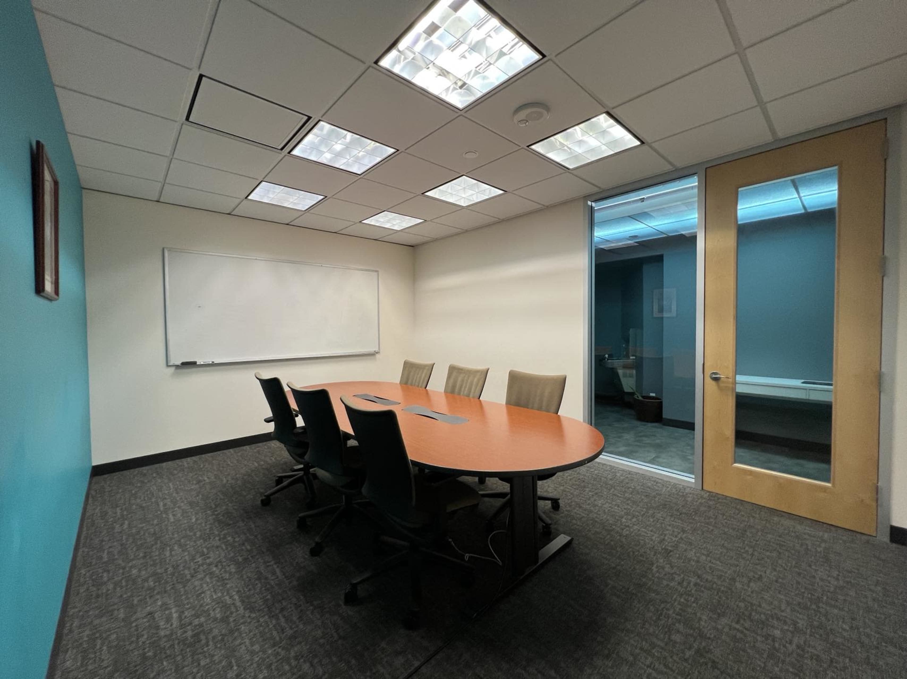 A conference room features a wooden table surrounded by chairs, a whiteboard on the wall, and a glass door leading to another area.