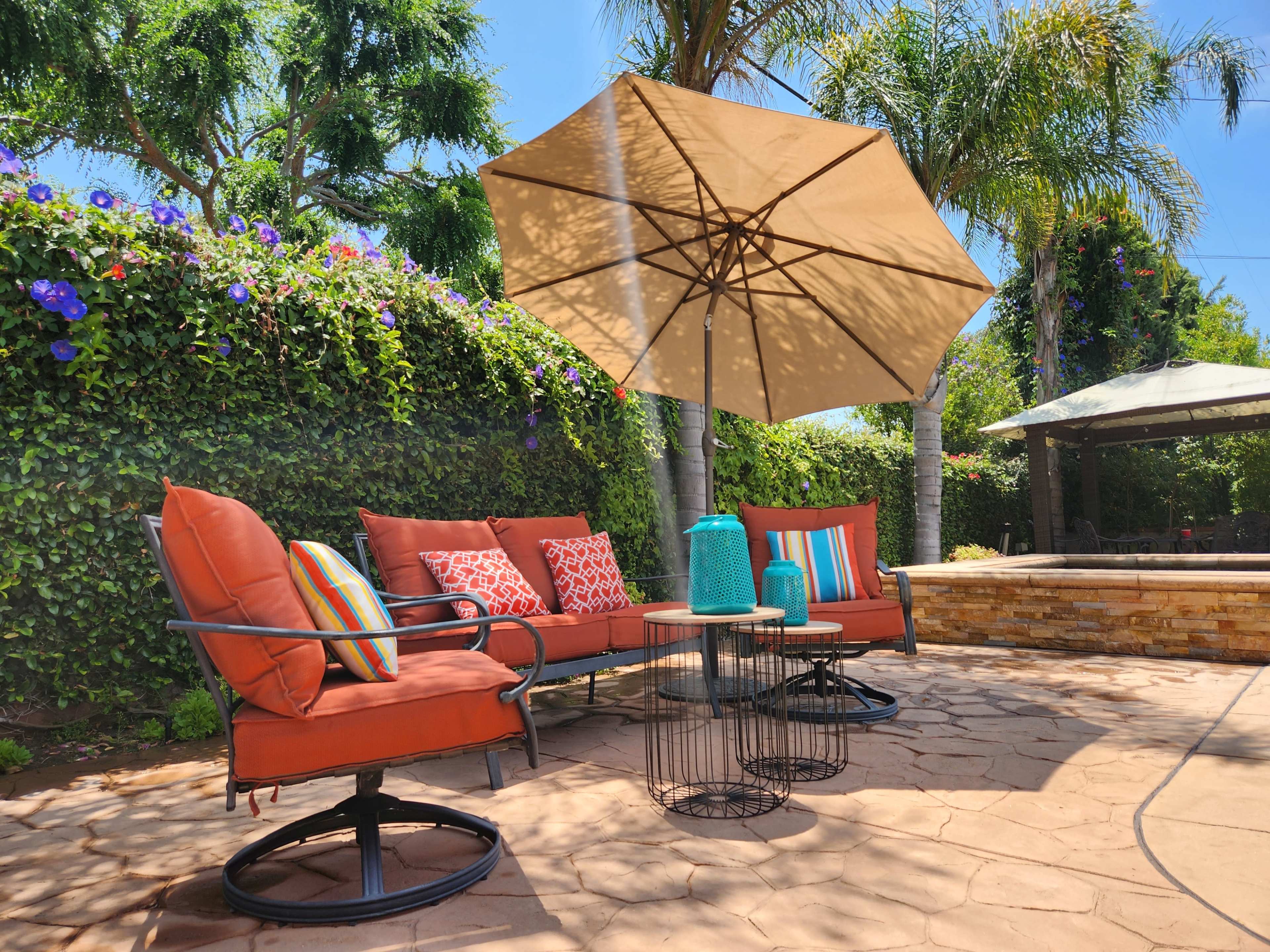 A patio area features a seating arrangement with orange cushions, a round table, and an umbrella, surrounded by greenery and flowering plants.