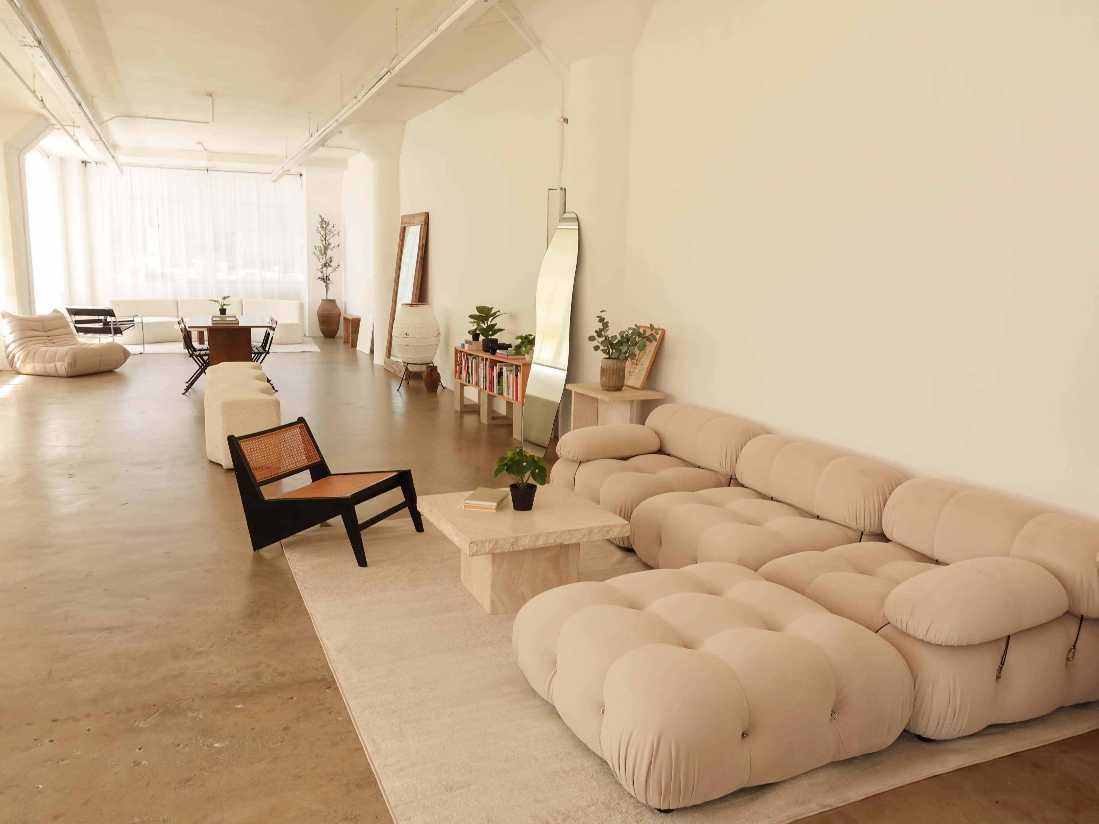 The image shows a spacious, minimalist interior featuring a large beige sectional sofa, a black wooden chair, a light-colored coffee table, and potted plants in a well-lit room.