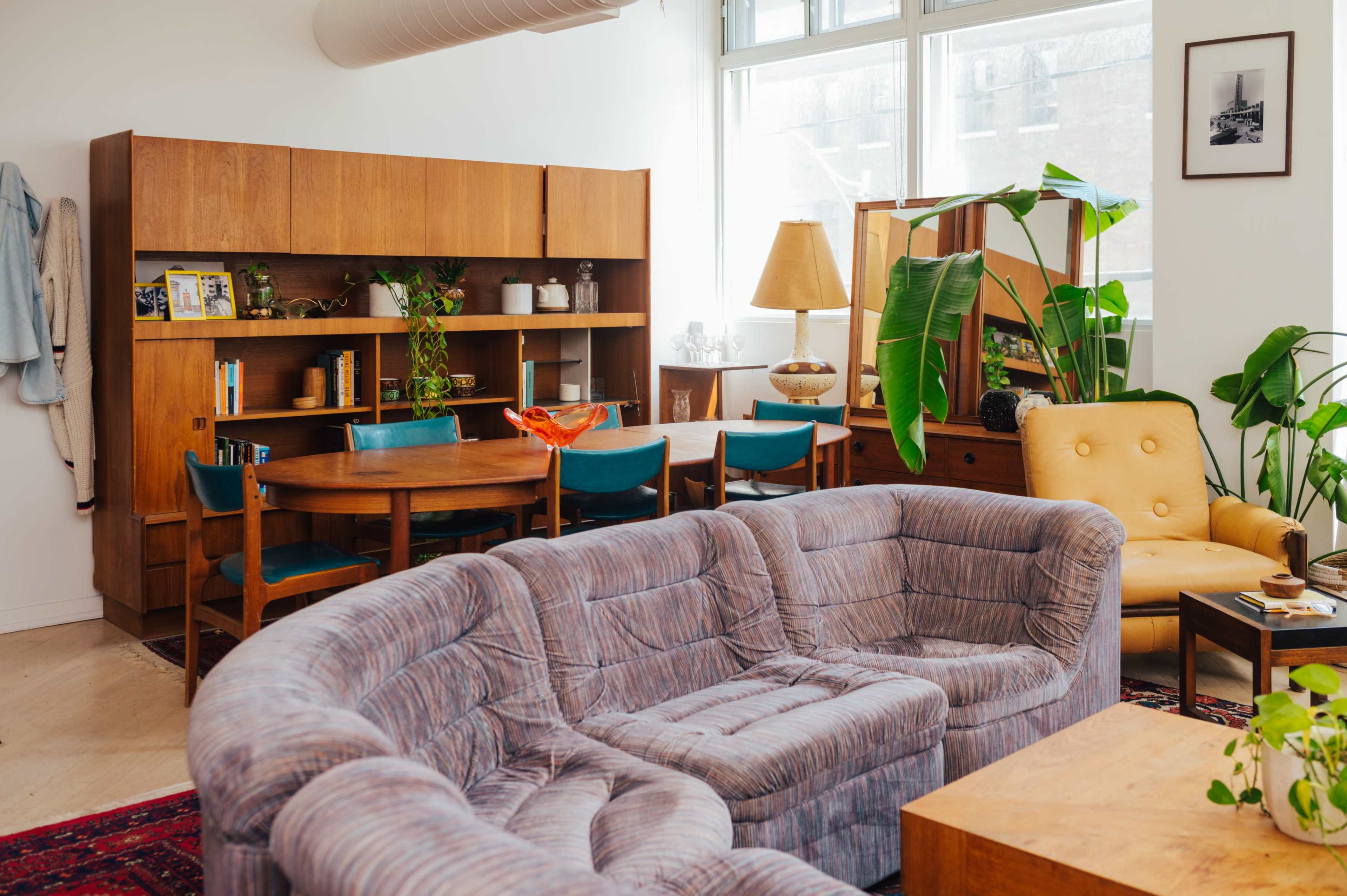 The image shows a modern living space featuring a curved gray sofa, a wooden dining table surrounded by chairs, and various houseplants.