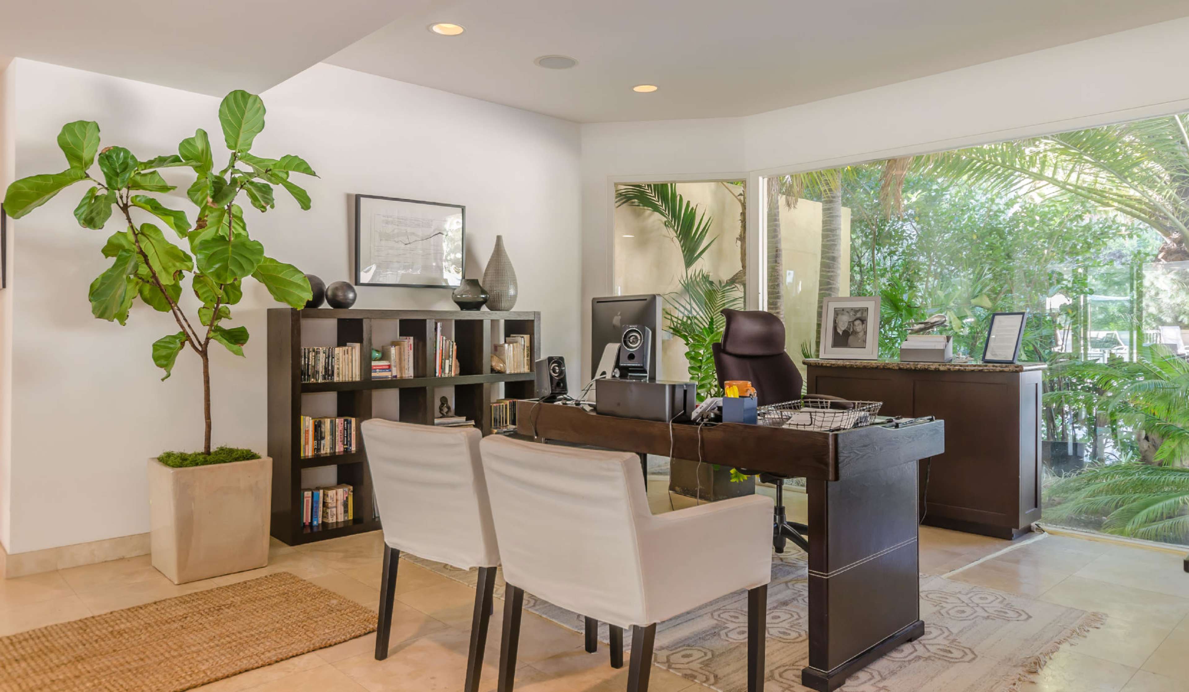 A modern home office features a desk with two chairs, a bookshelf filled with books, and large windows overlooking greenery.