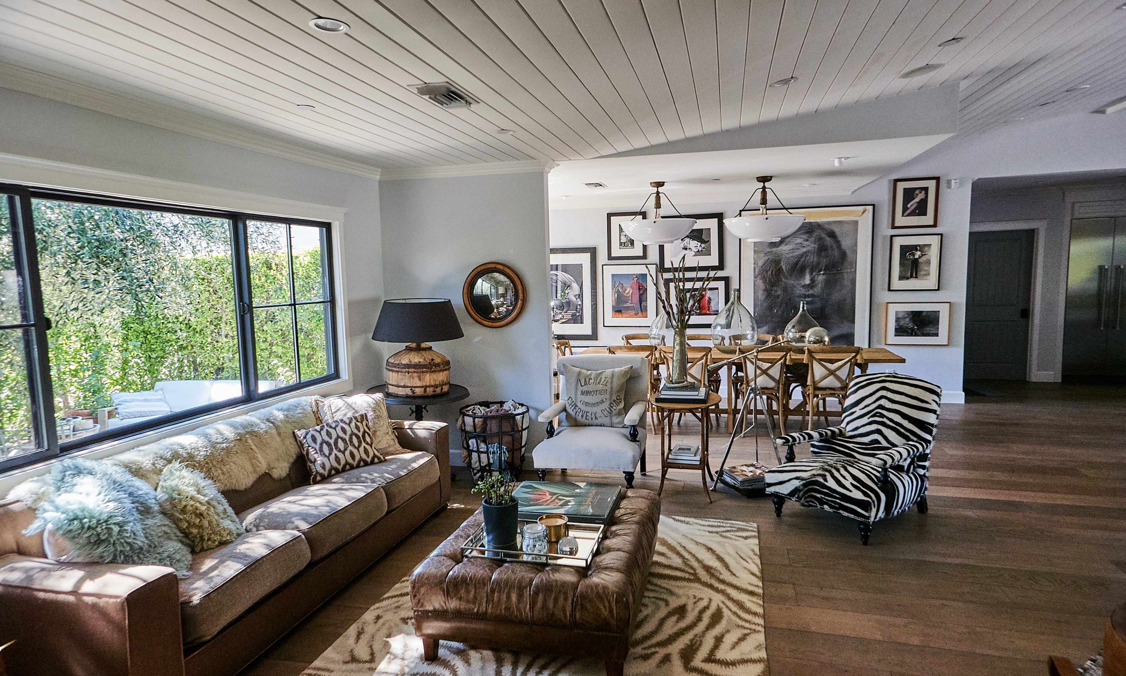 The image shows a modern living room with a leather sofa, a glass coffee table, and a dining area featuring a wooden table surrounded by chairs, decorated with various artworks on the walls.