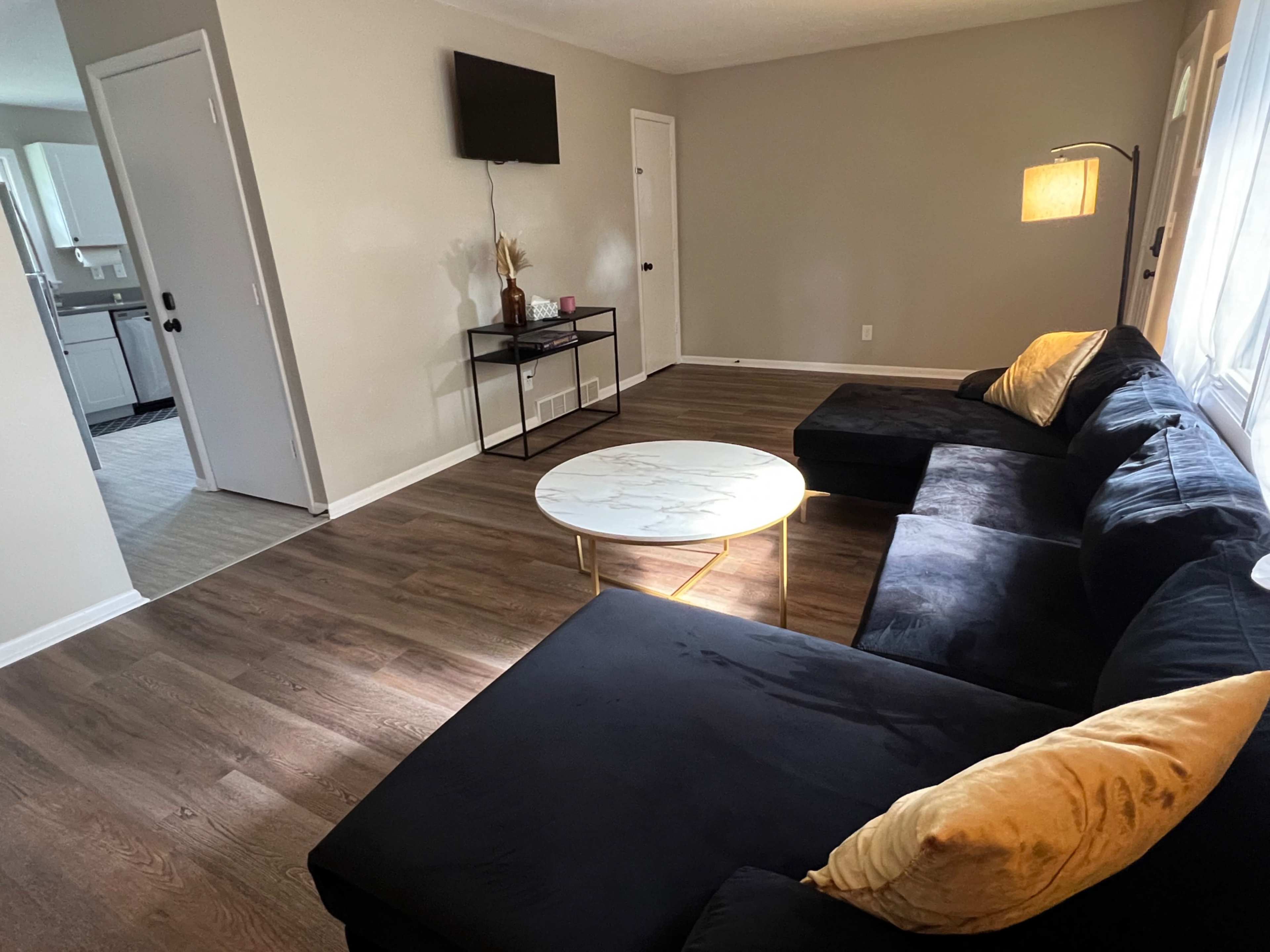 A living room features a black sectional sofa, a marble coffee table, a wall-mounted television, and a doorway leading to a kitchen.