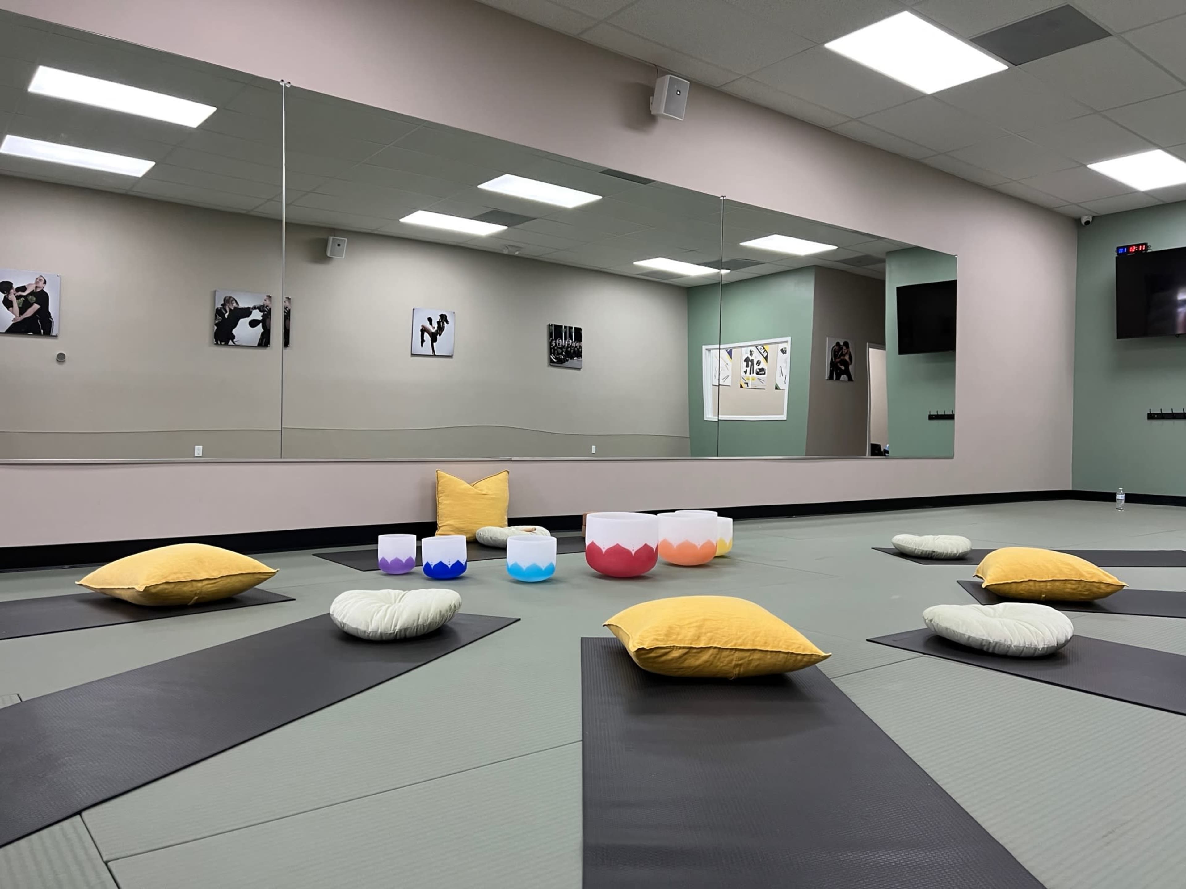 A calm studio space is arranged with mats, cushions, and a center display, featuring mirrored walls and artwork.