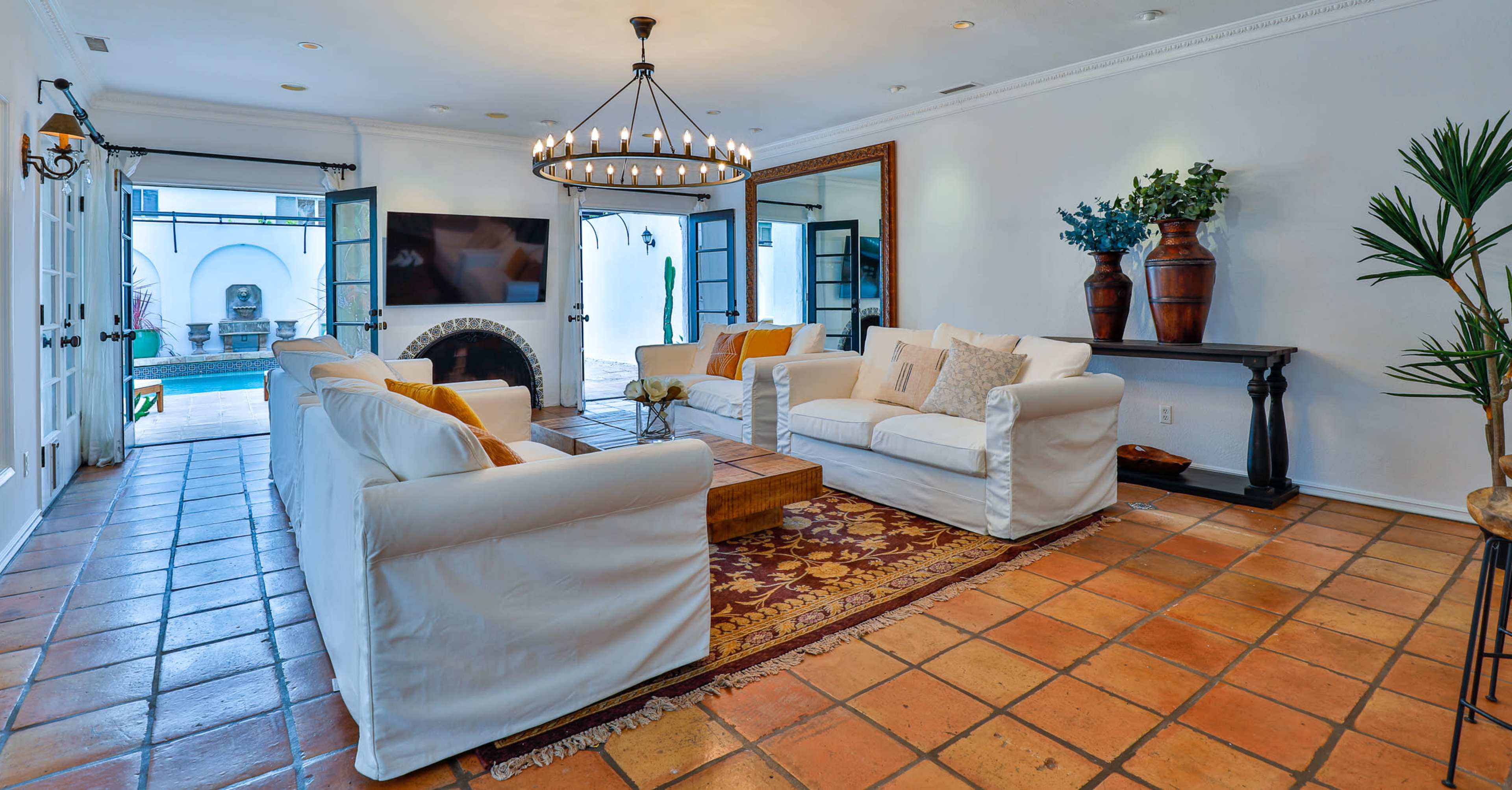 The image shows a living room with white sofas, a wooden coffee table on a patterned rug, and a large television mounted on the wall, all illuminated by a circular chandelier.