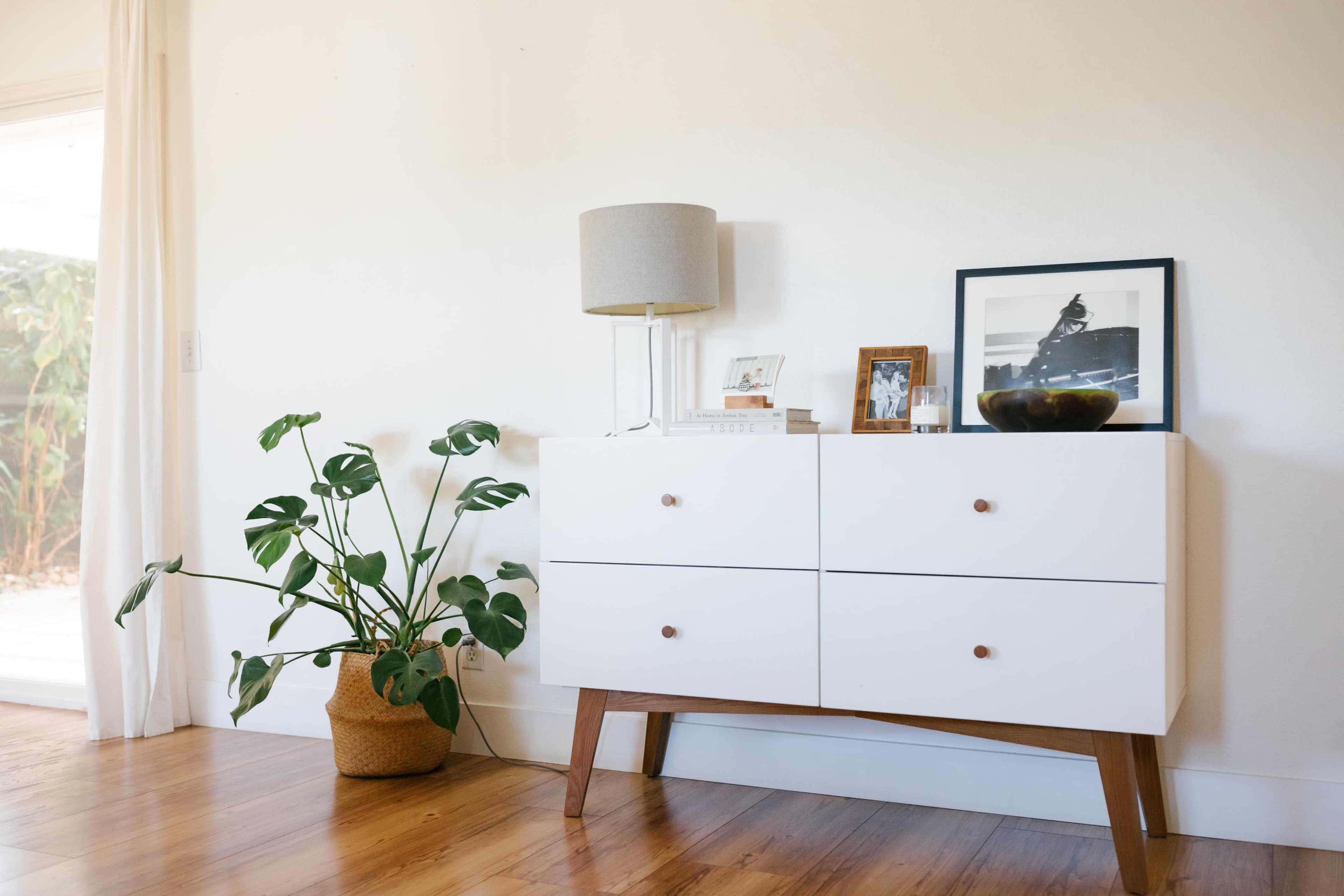A modern white sideboard with wooden legs, a lamp, framed photos, and a plant sits against a wall in a bright room.