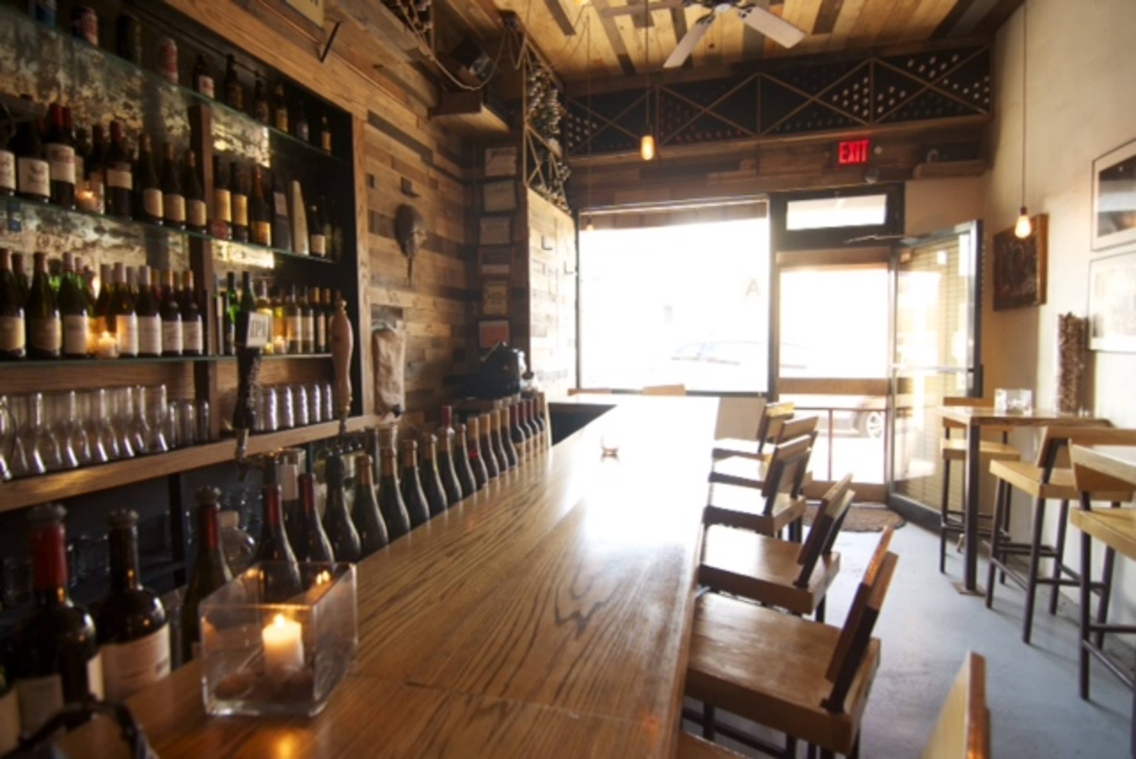 The interior of a restaurant or bar with a wooden counter, shelves of wine bottles, and a seating area with wooden stools and tables.