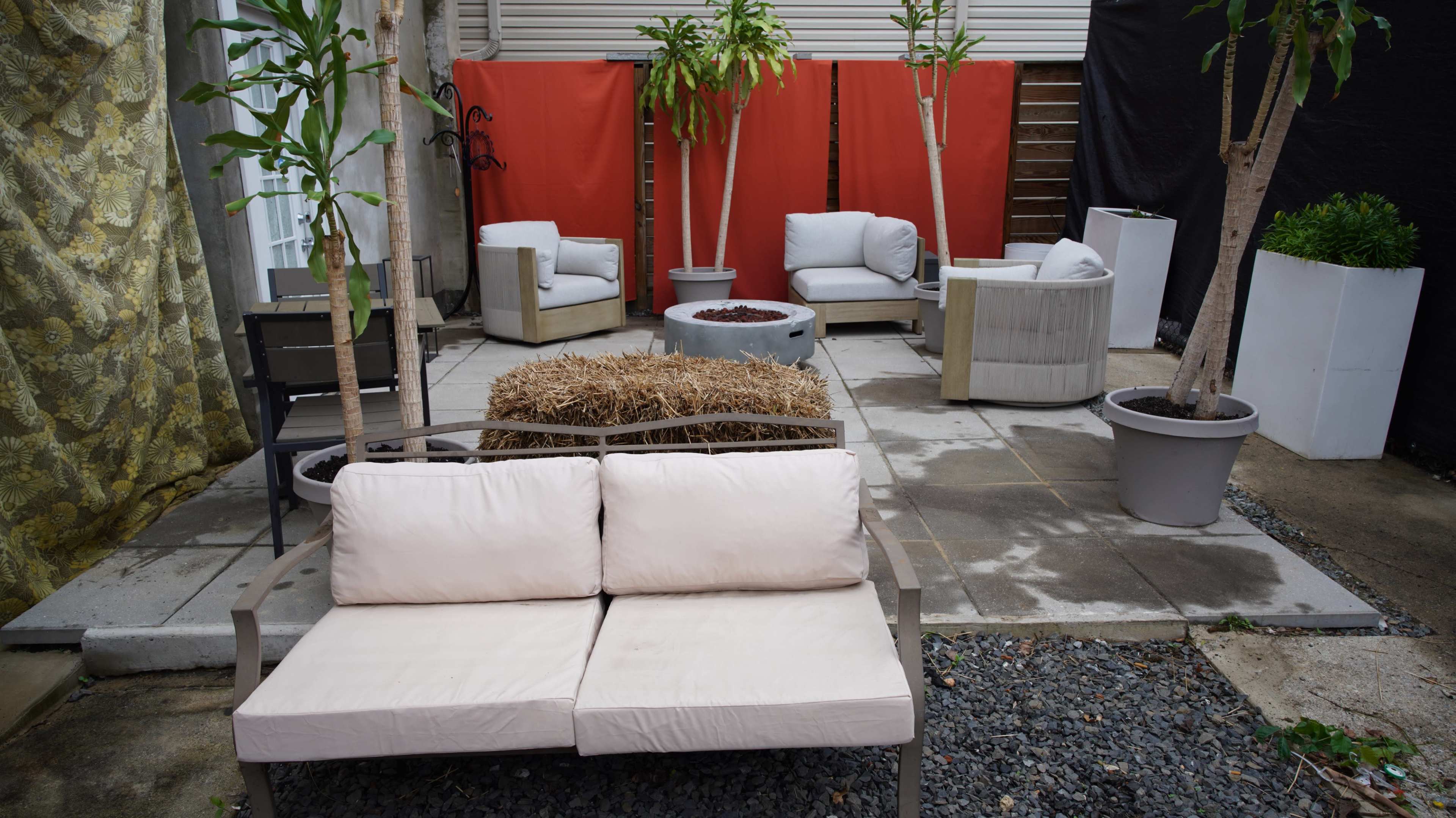 The image shows an outdoor seating area with several chairs and tables arranged around a fire pit, surrounded by greenery and fabric dividers.