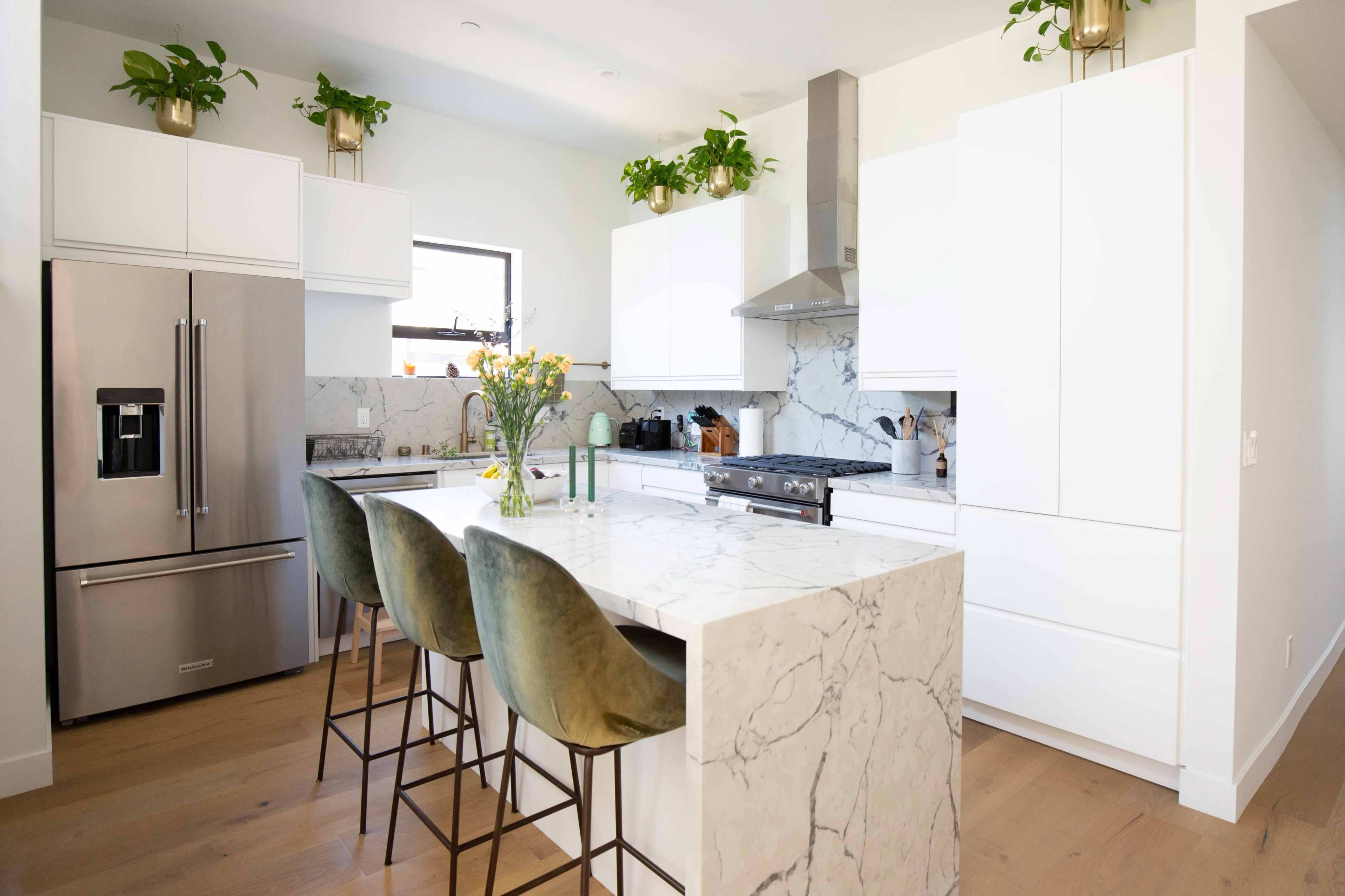A modern kitchen features a marble island with four green bar stools, stainless steel appliances, and white cabinetry adorned with potted plants.