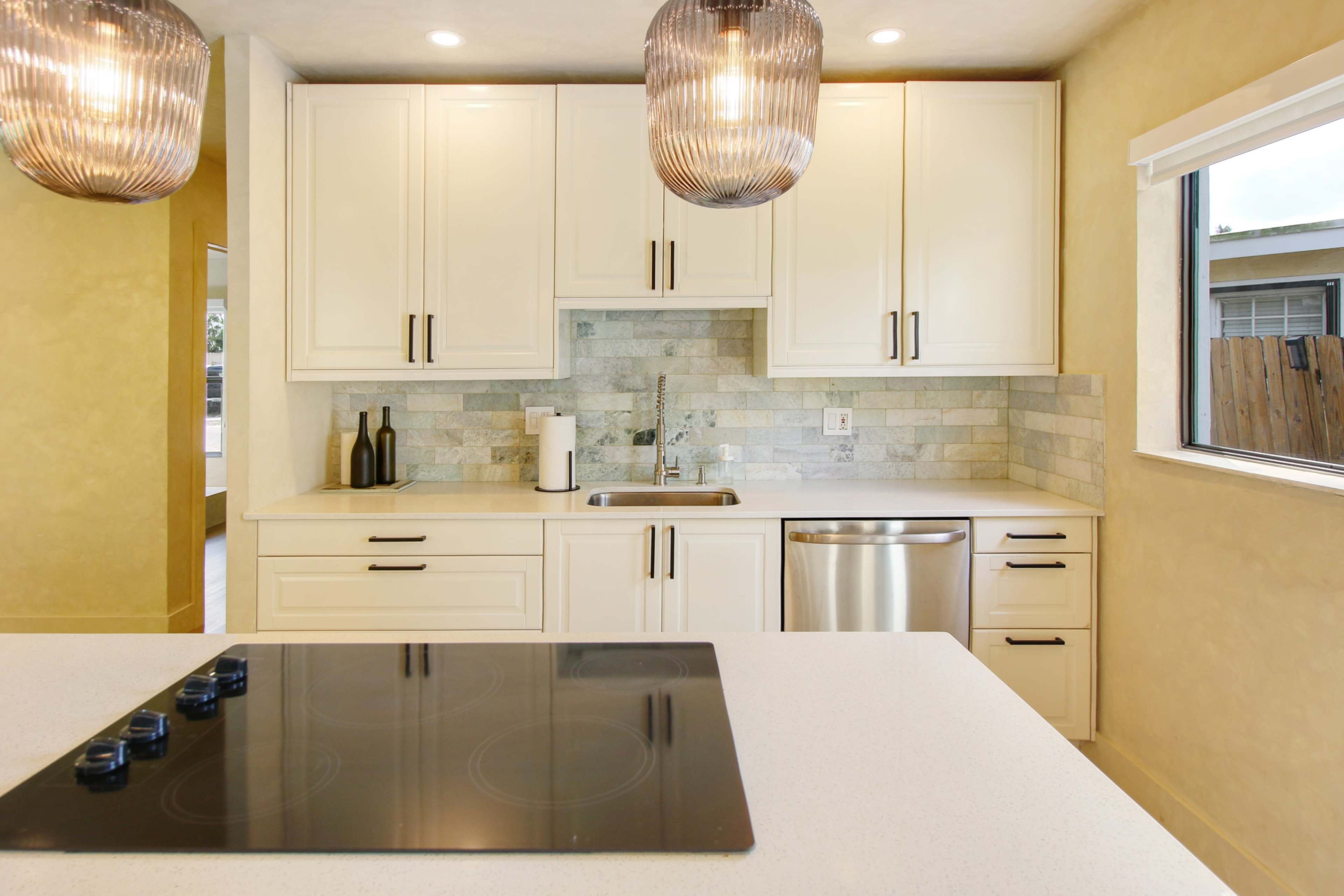 A modern kitchen features white cabinetry, a stainless steel sink, and an island with a built-in cooktop, illuminated by pendant lights.