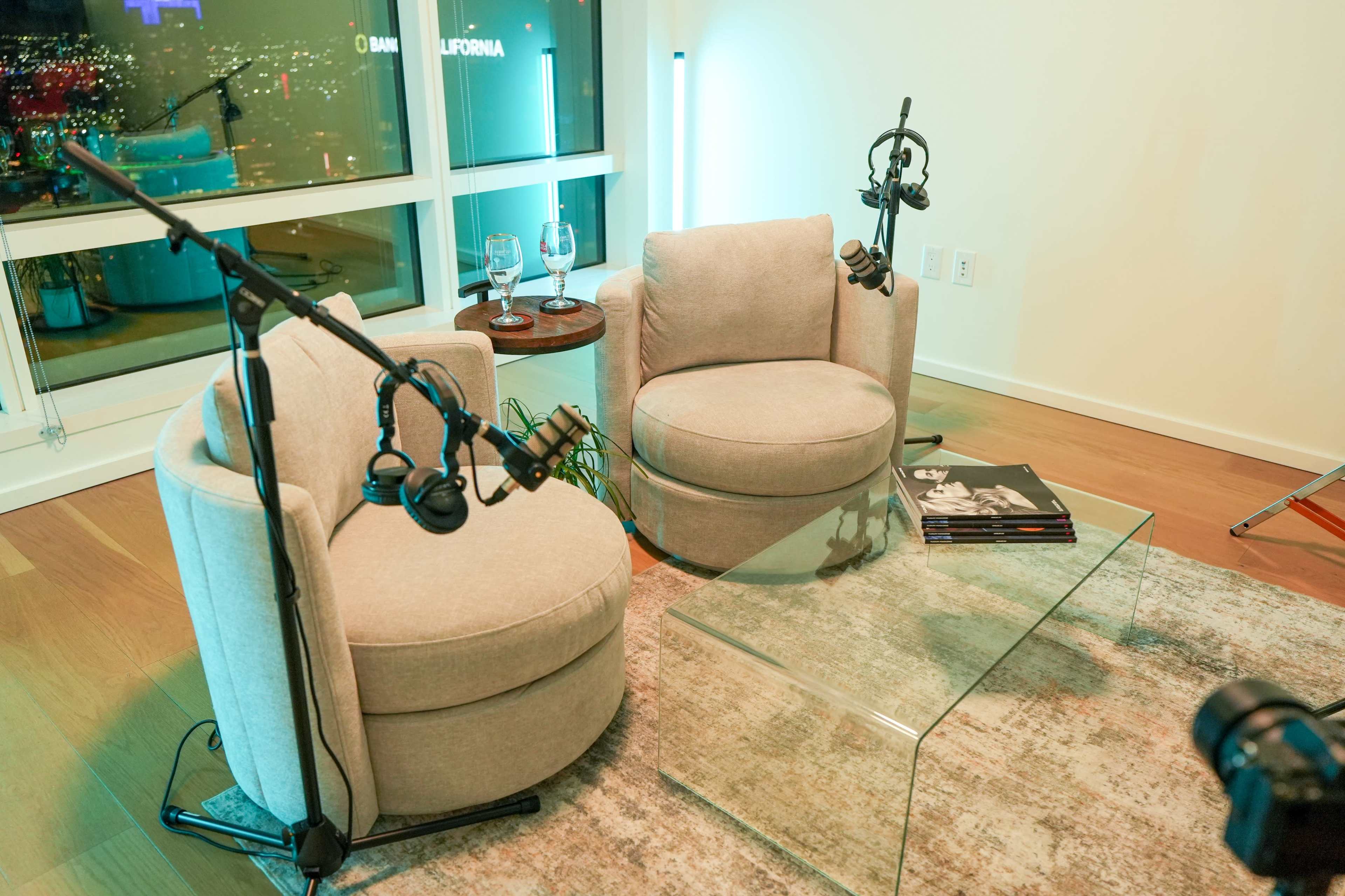 The scene features two upholstered chairs positioned around a glass coffee table, with microphones mounted on stands nearby, set against a backdrop of city lights visible through large windows.