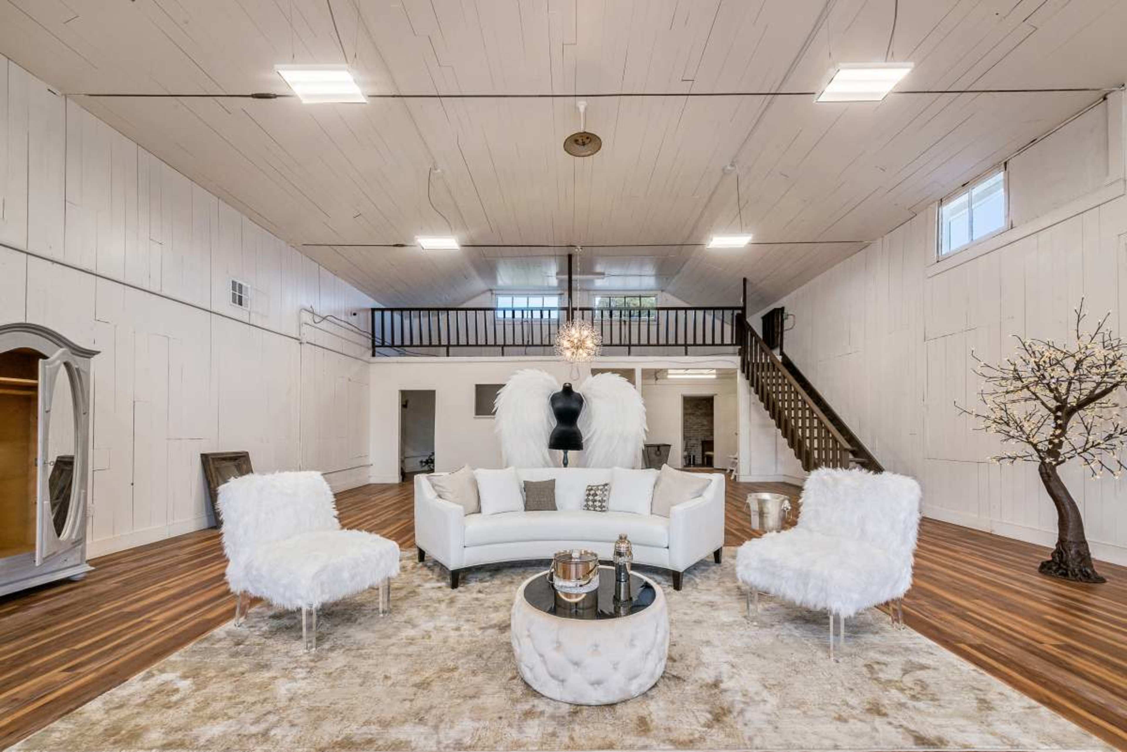 The image shows a spacious, well-lit room featuring a white sectional sofa, two fluffy chairs, a round coffee table, and minimalist decor against wooden flooring and white walls.