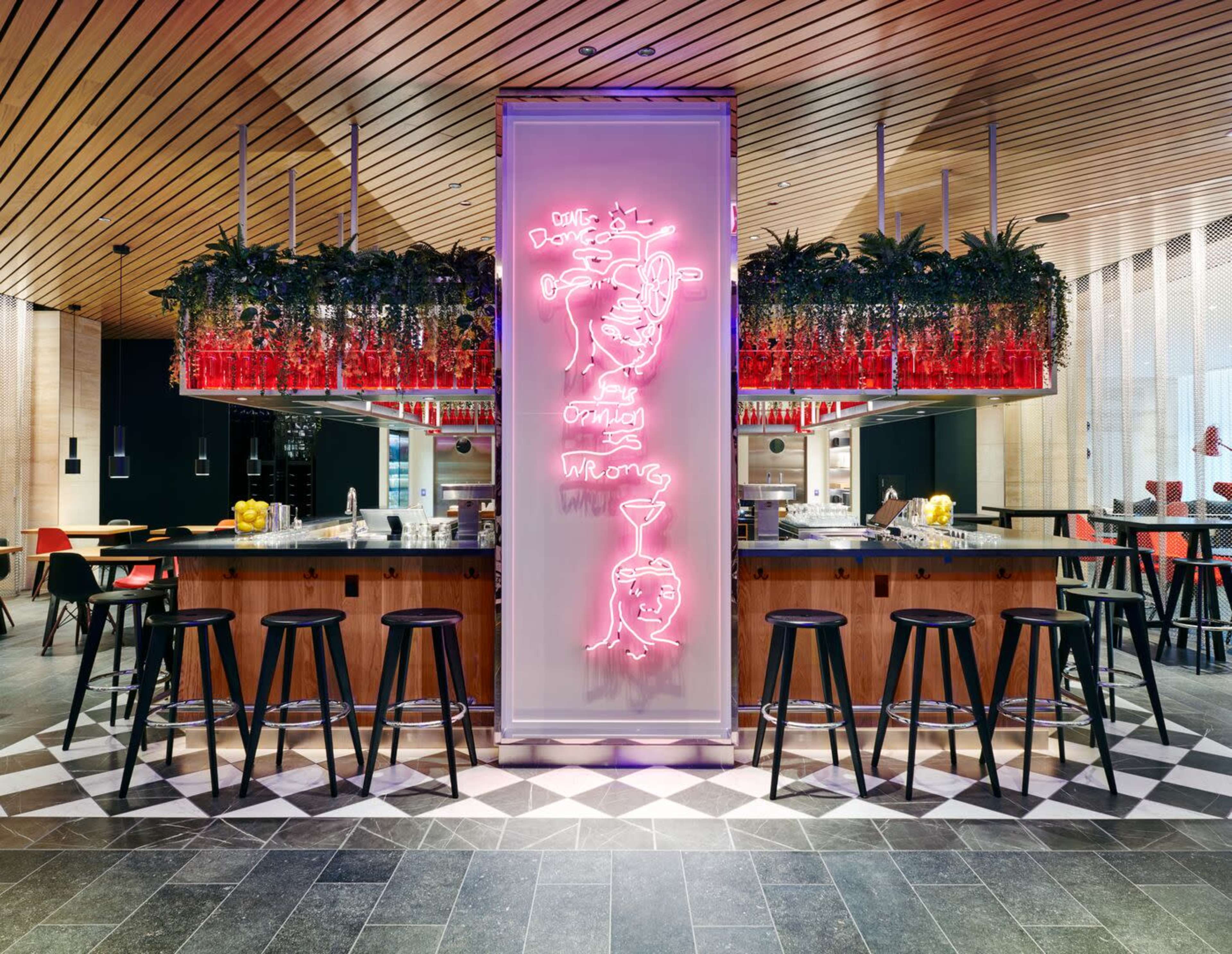 A modern bar features a neon sign and is surrounded by high stools, with decorative plants hanging above and a patterned floor.