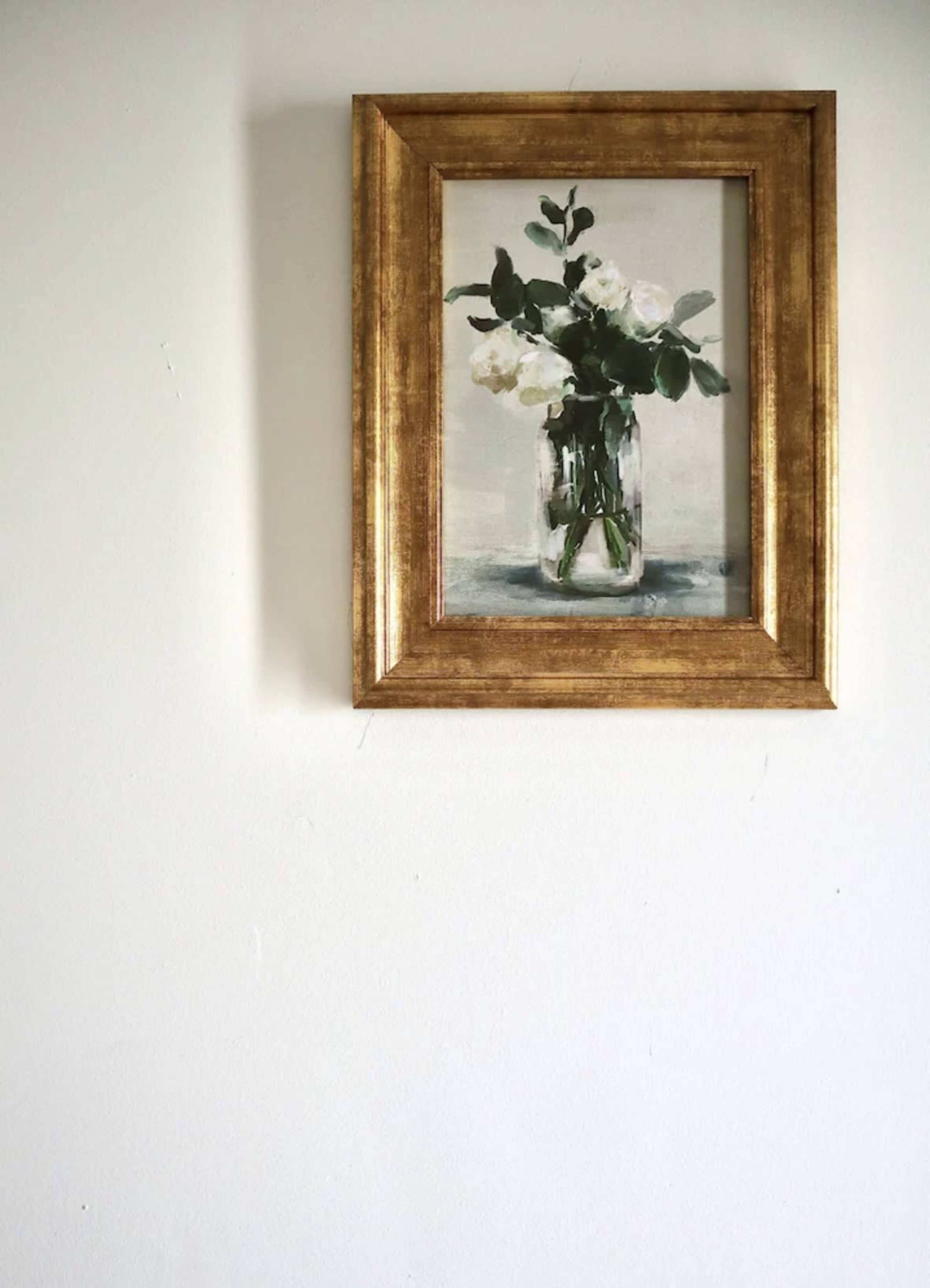 A gold-framed painting of white flowers in a glass vase is hung on a plain white wall.