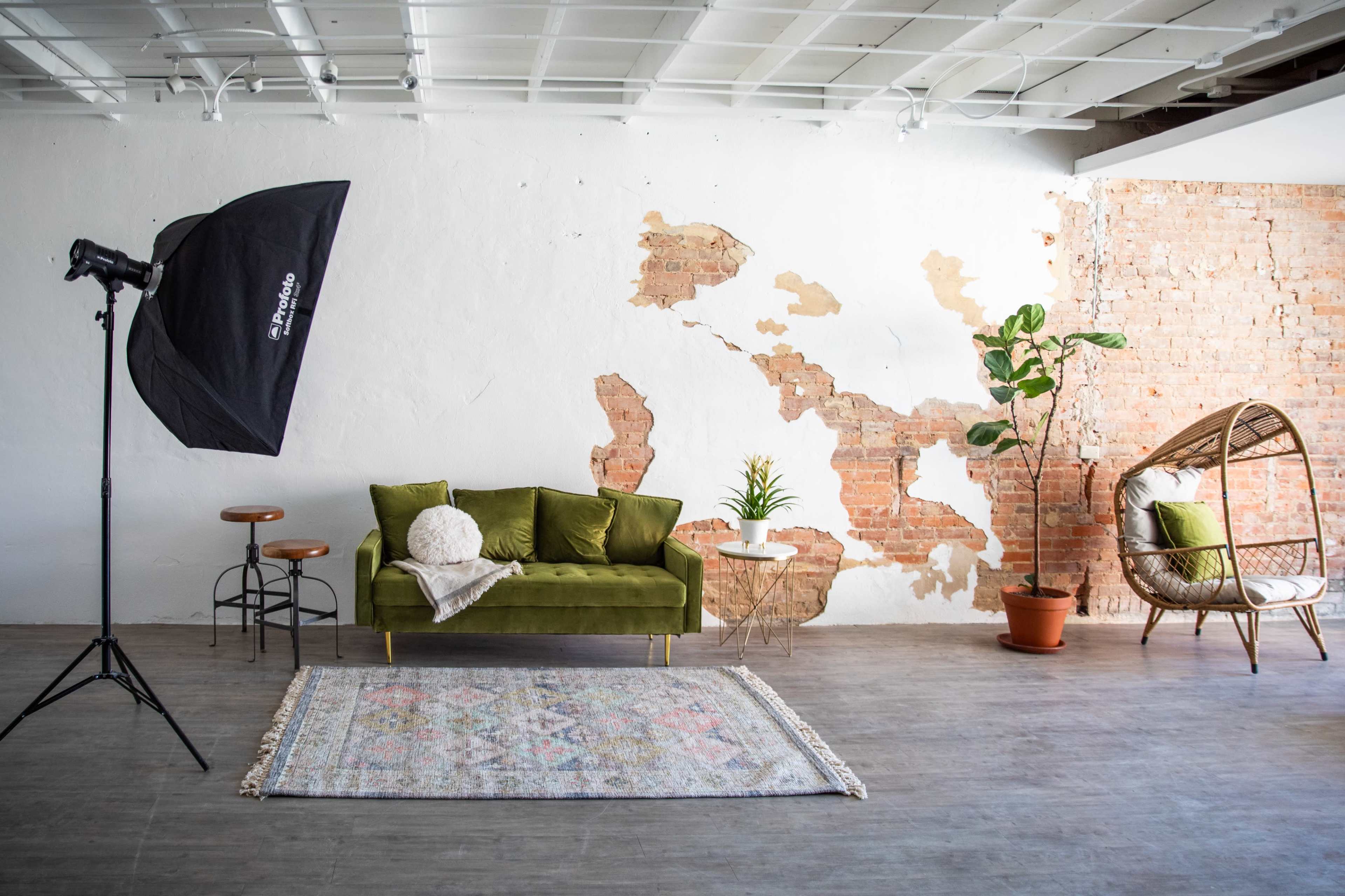 The scene features a well-lit room with a green sofa, a round rug, a potted plant, and a wicker chair against a wall with exposed brick and a lighting setup.