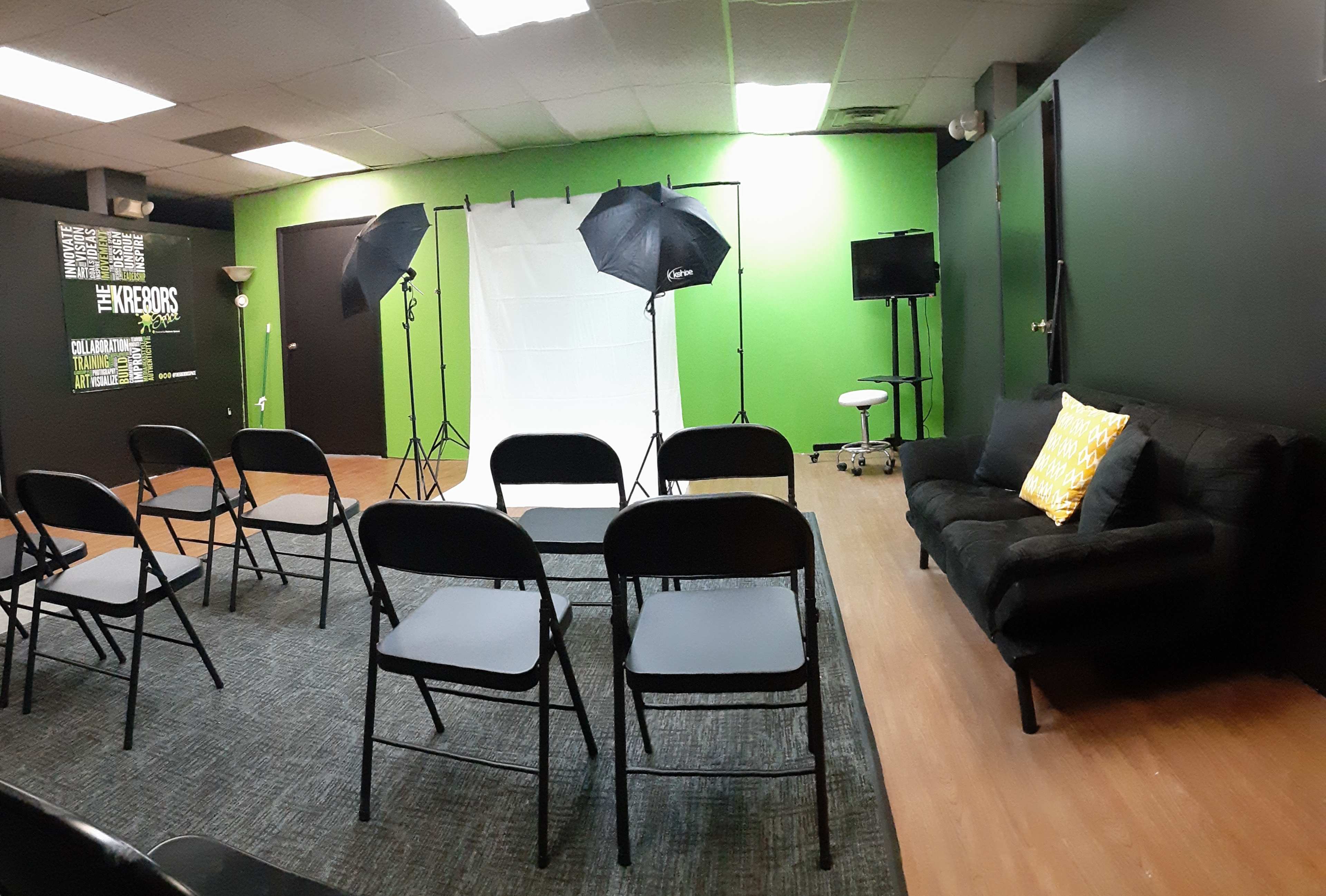 A small room set up for an event, featuring folding chairs arranged in rows, a couch, lighting equipment, and a green accent wall.