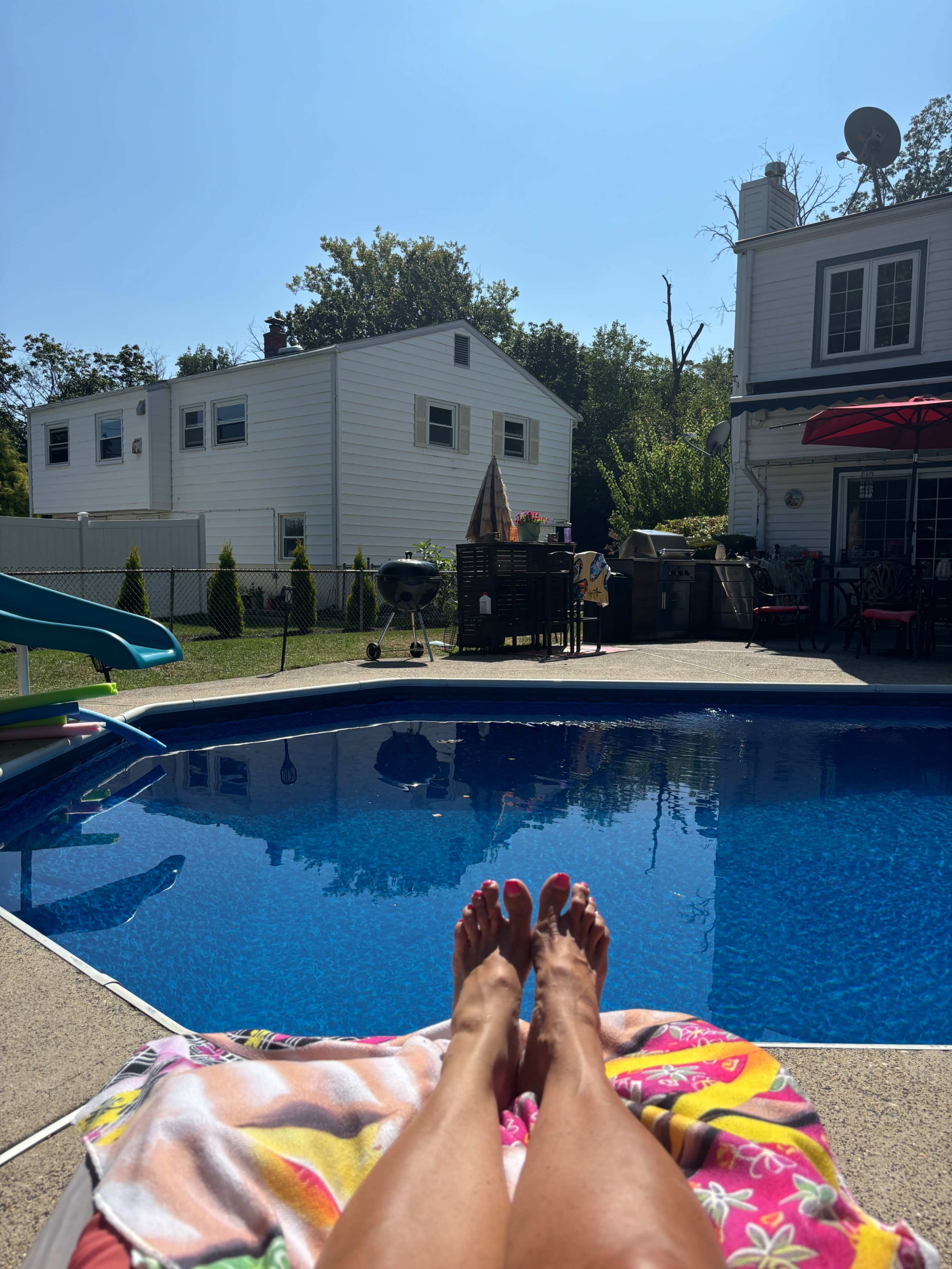A pair of bare feet rests on a colorful towel beside a clear pool, with houses and greenery visible in the background.