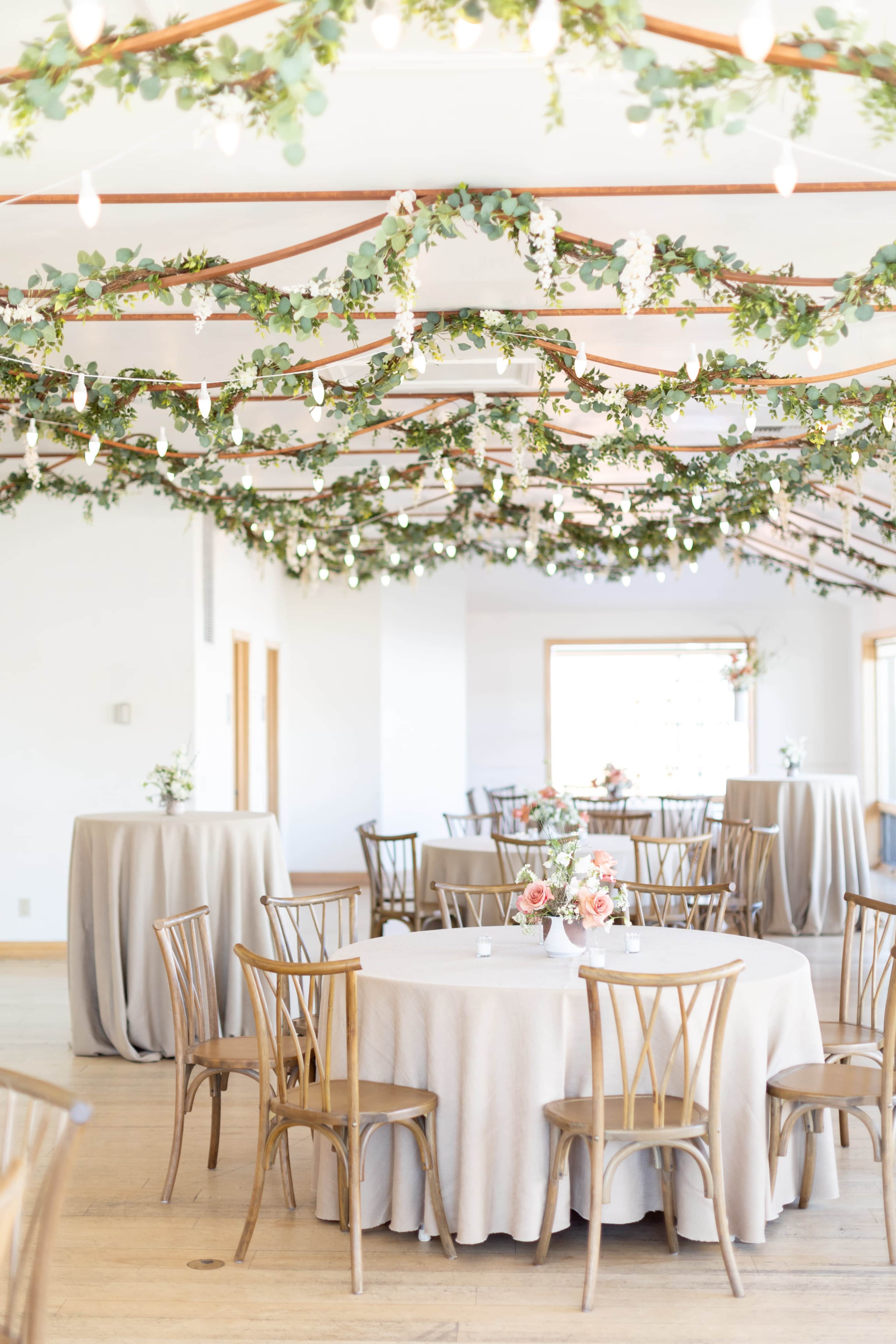 The image shows a banquet hall decorated with greenery and string lights, featuring round tables covered with light fabric.