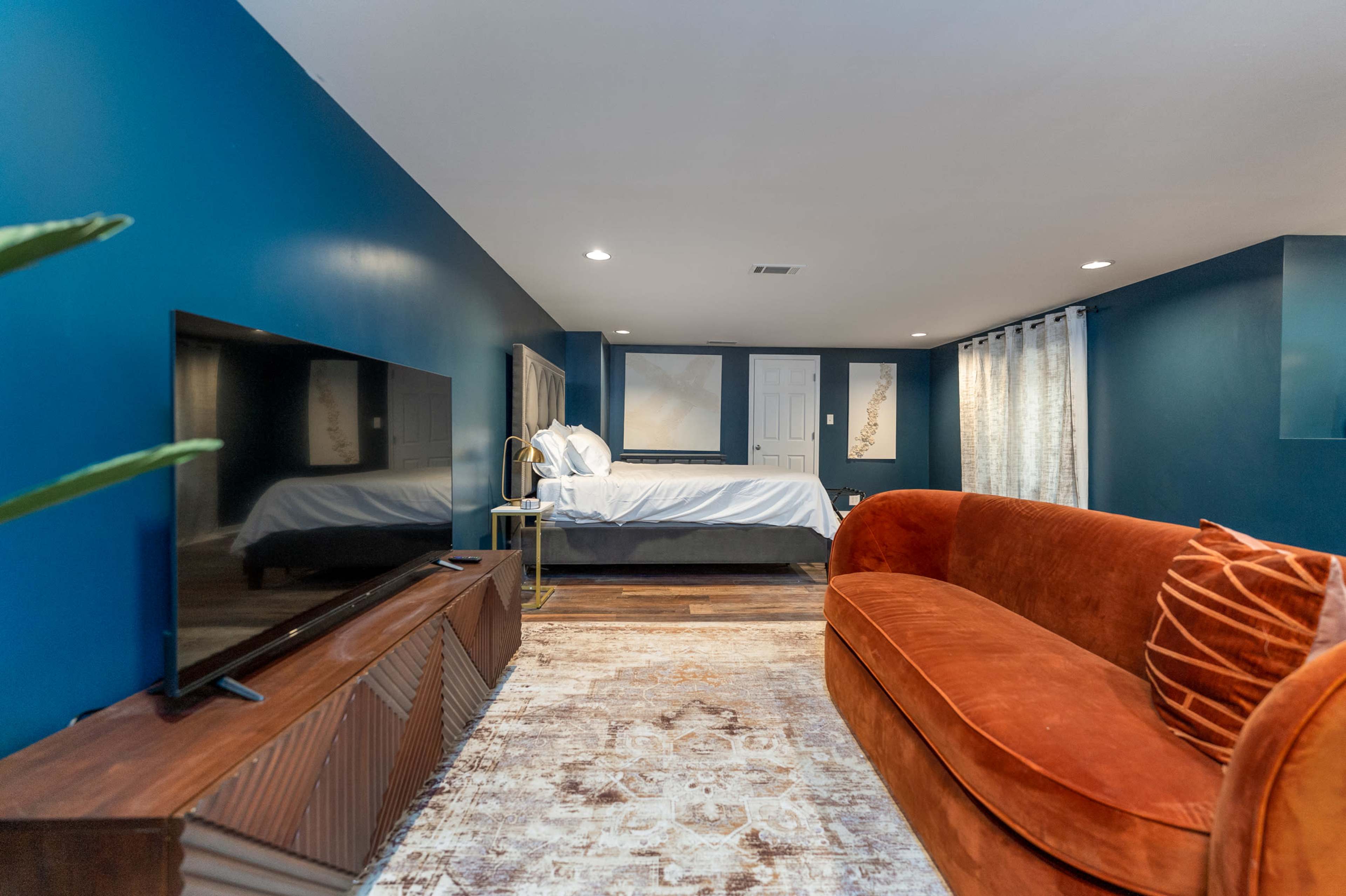 A modern interior of a room featuring a bed, a large television on a wooden stand, and a curved orange couch against a dark blue wall.
