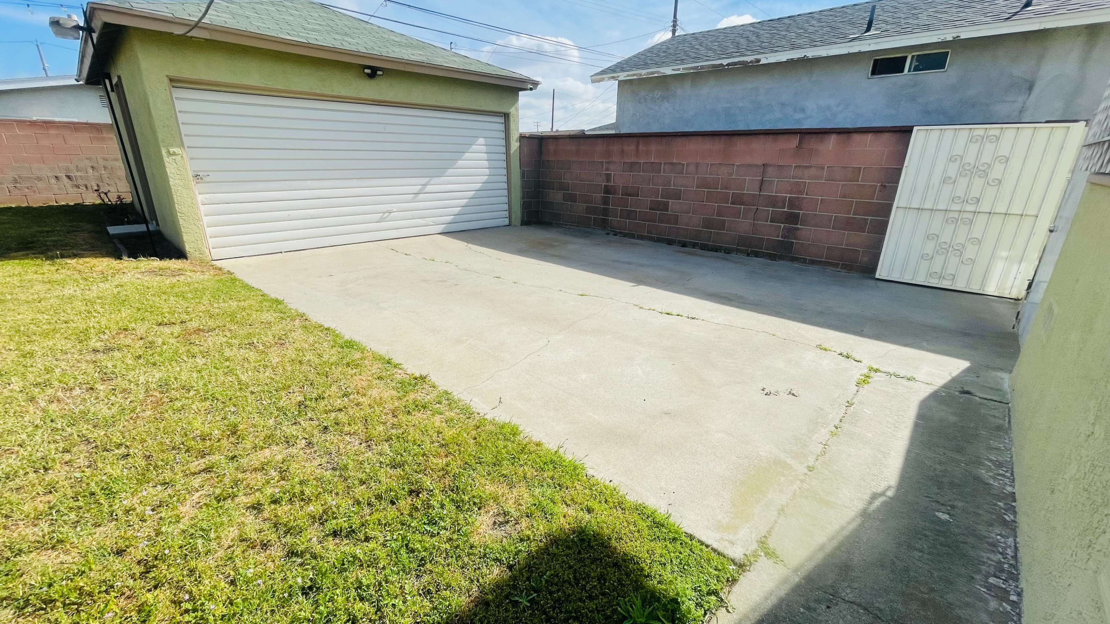 The image shows a vacant concrete driveway leading to a garage with a closed white door, bordered by a brick wall and a gated entrance.