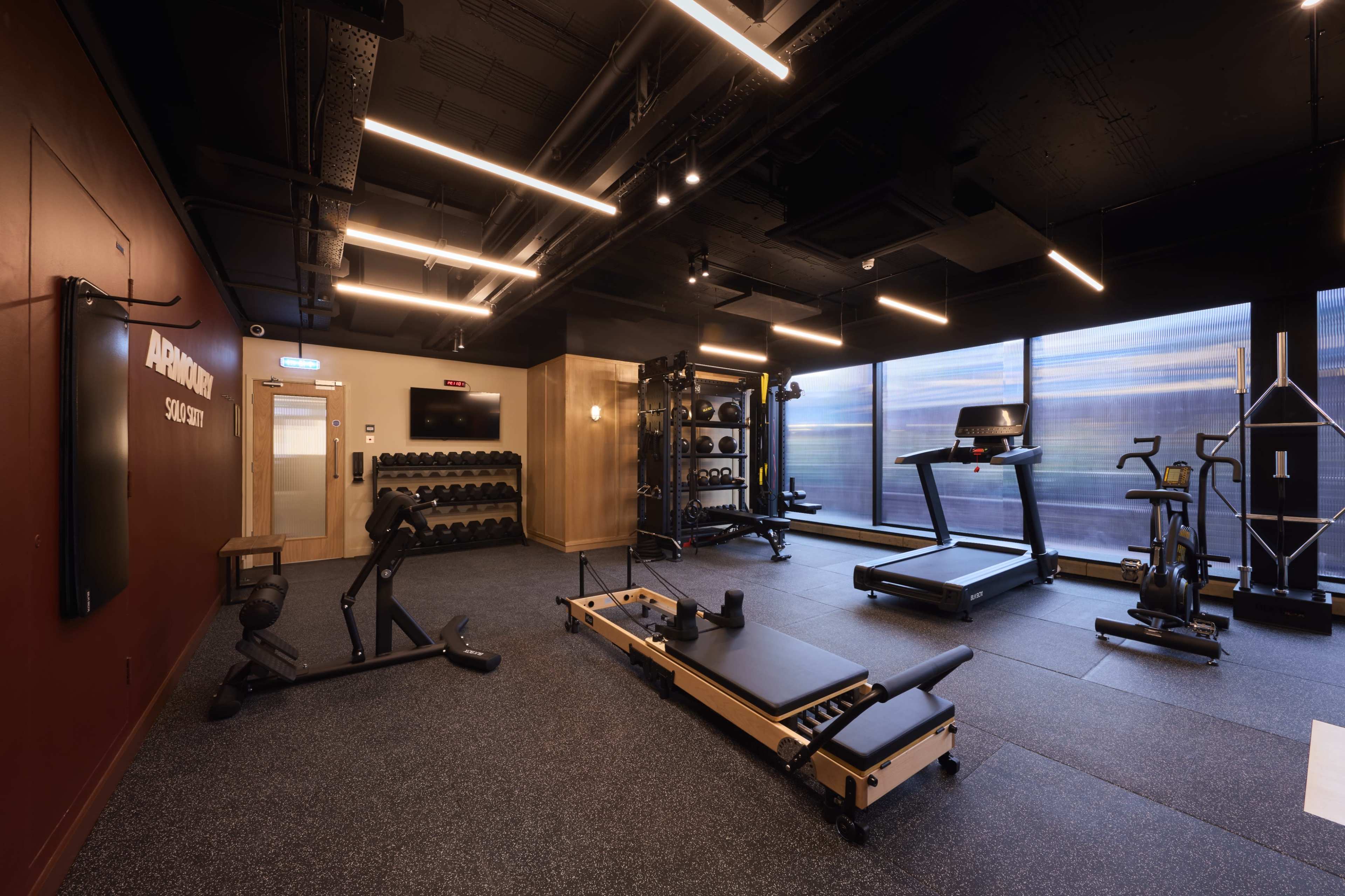 The image shows a modern gym interior with various fitness equipment, including weights, a treadmill, and a reformer, set against a backdrop of large windows.