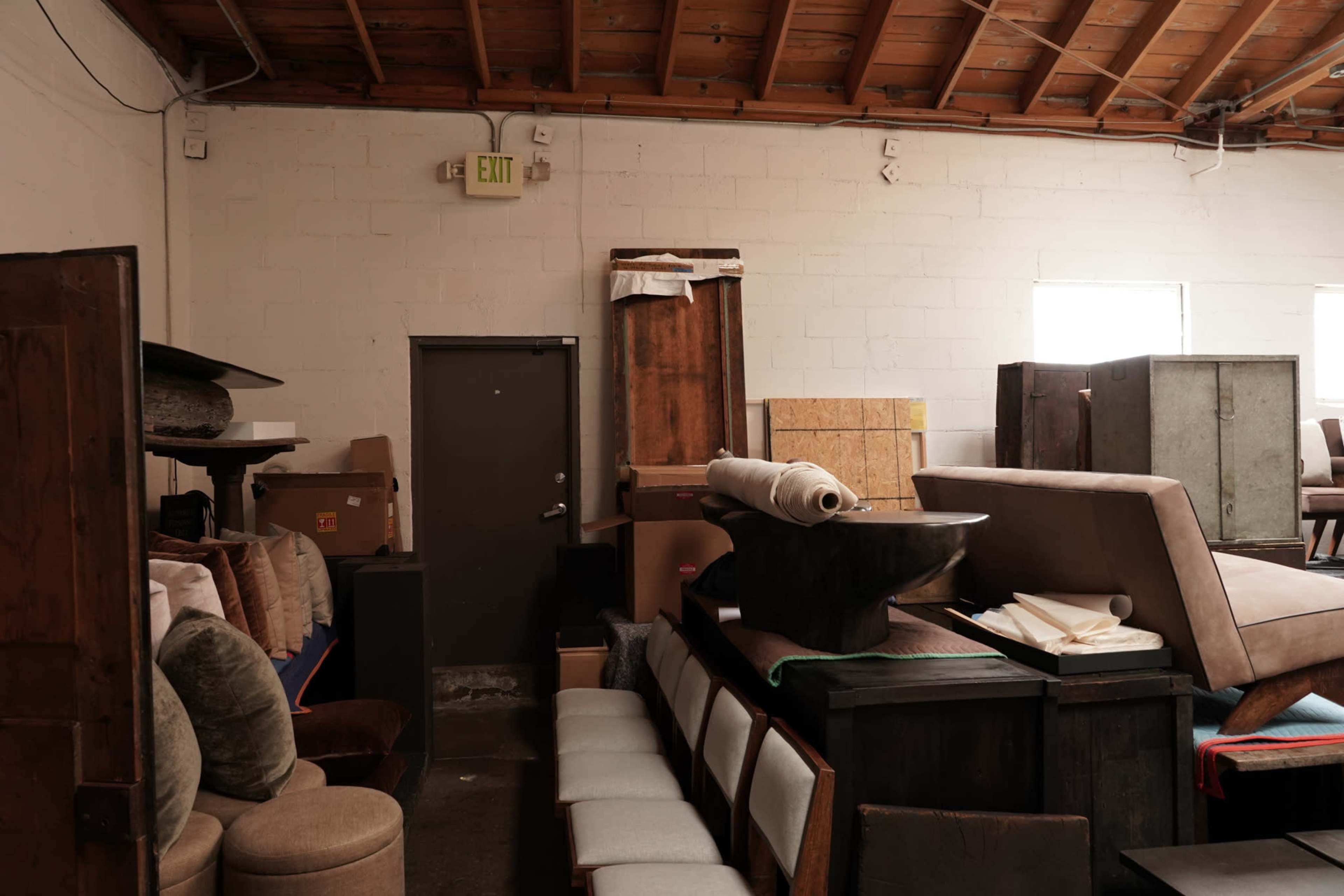 The image shows a cluttered warehouse space filled with various pieces of furniture and boxes stacked against the walls.