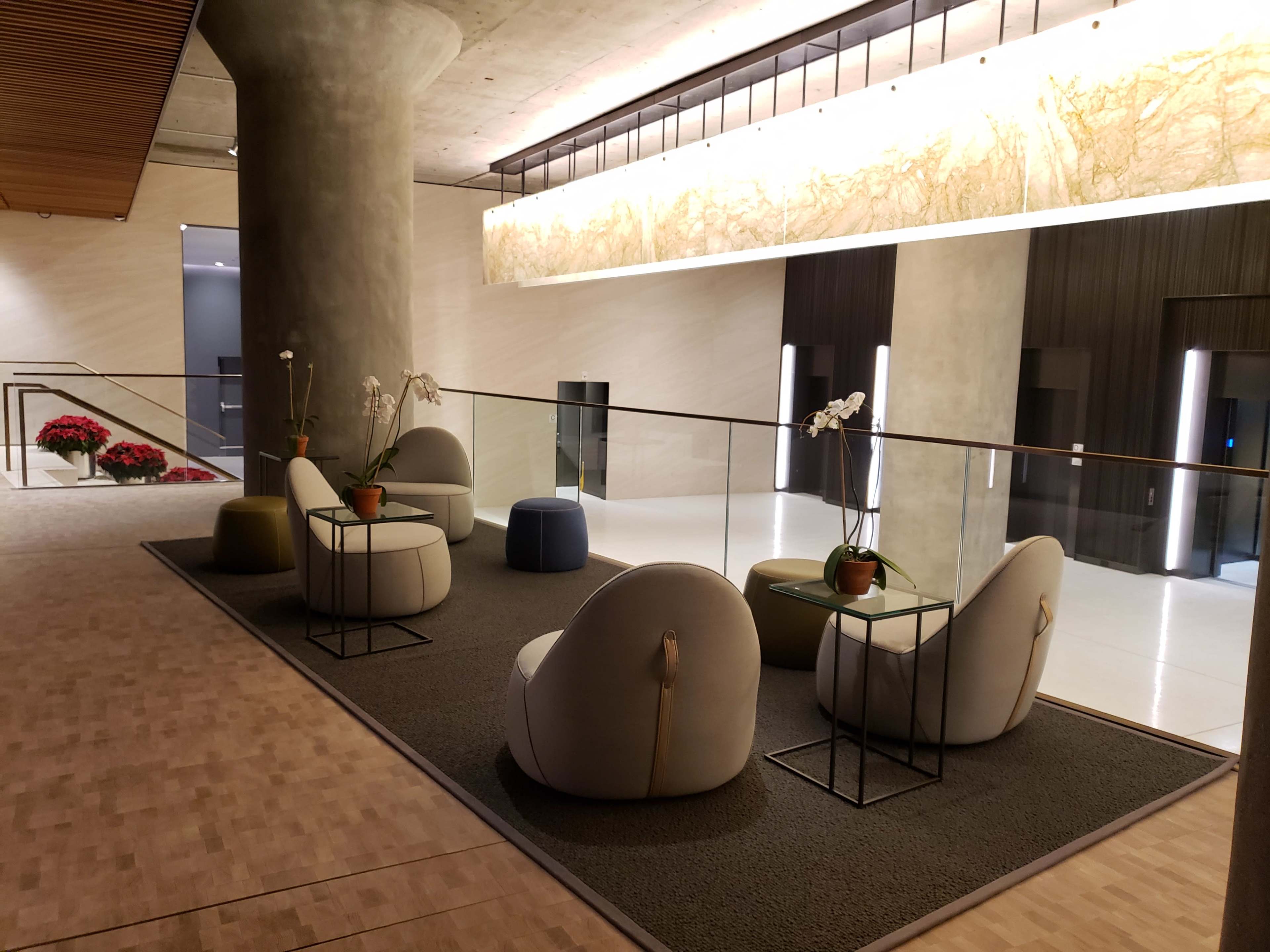 The image shows a modern lobby area with round seating, small tables, and decorative plants on a carpeted floor.