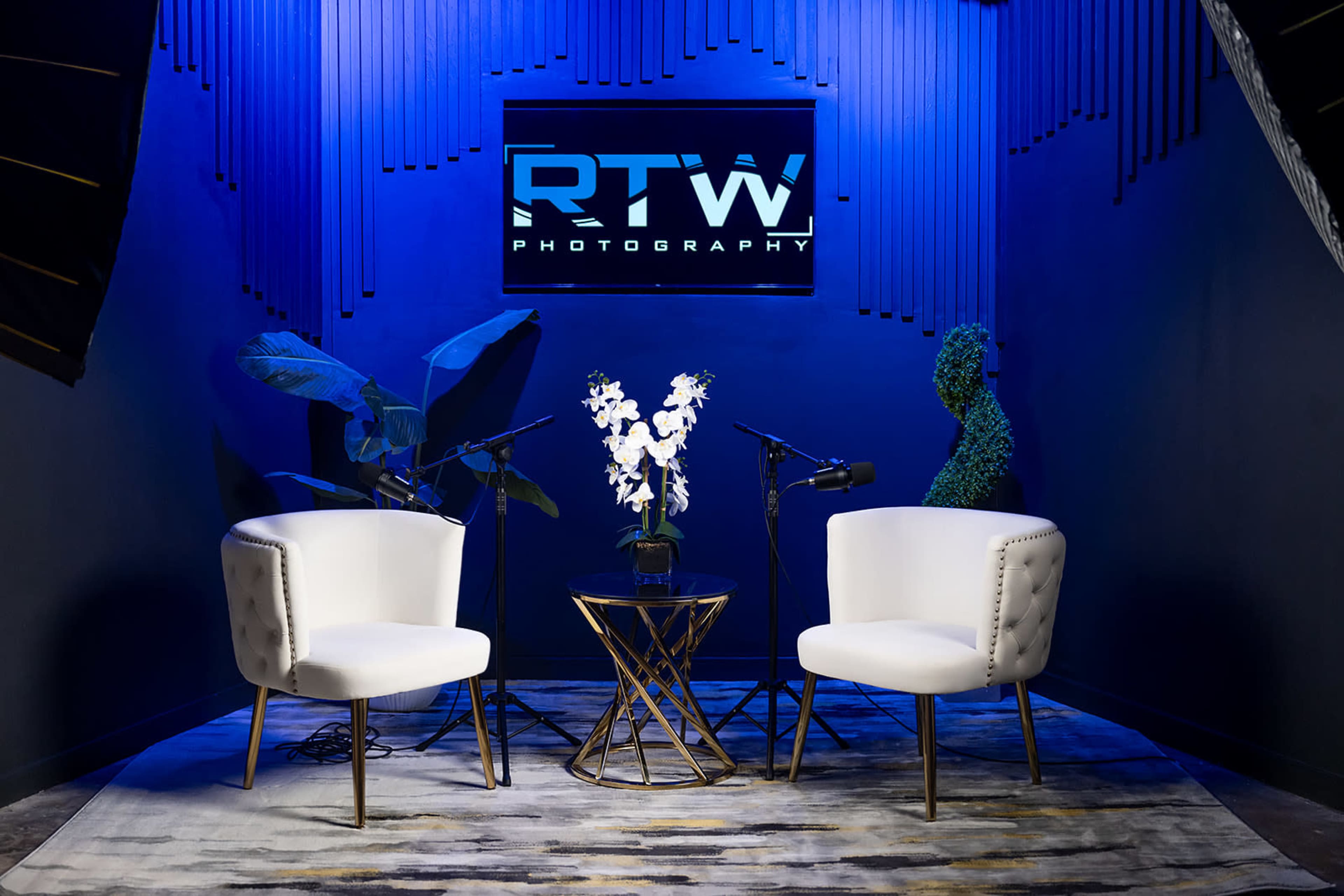 The image shows a modern photography studio setup with two white chairs, a small table with a floral arrangement, and microphones positioned for an interview or discussion.