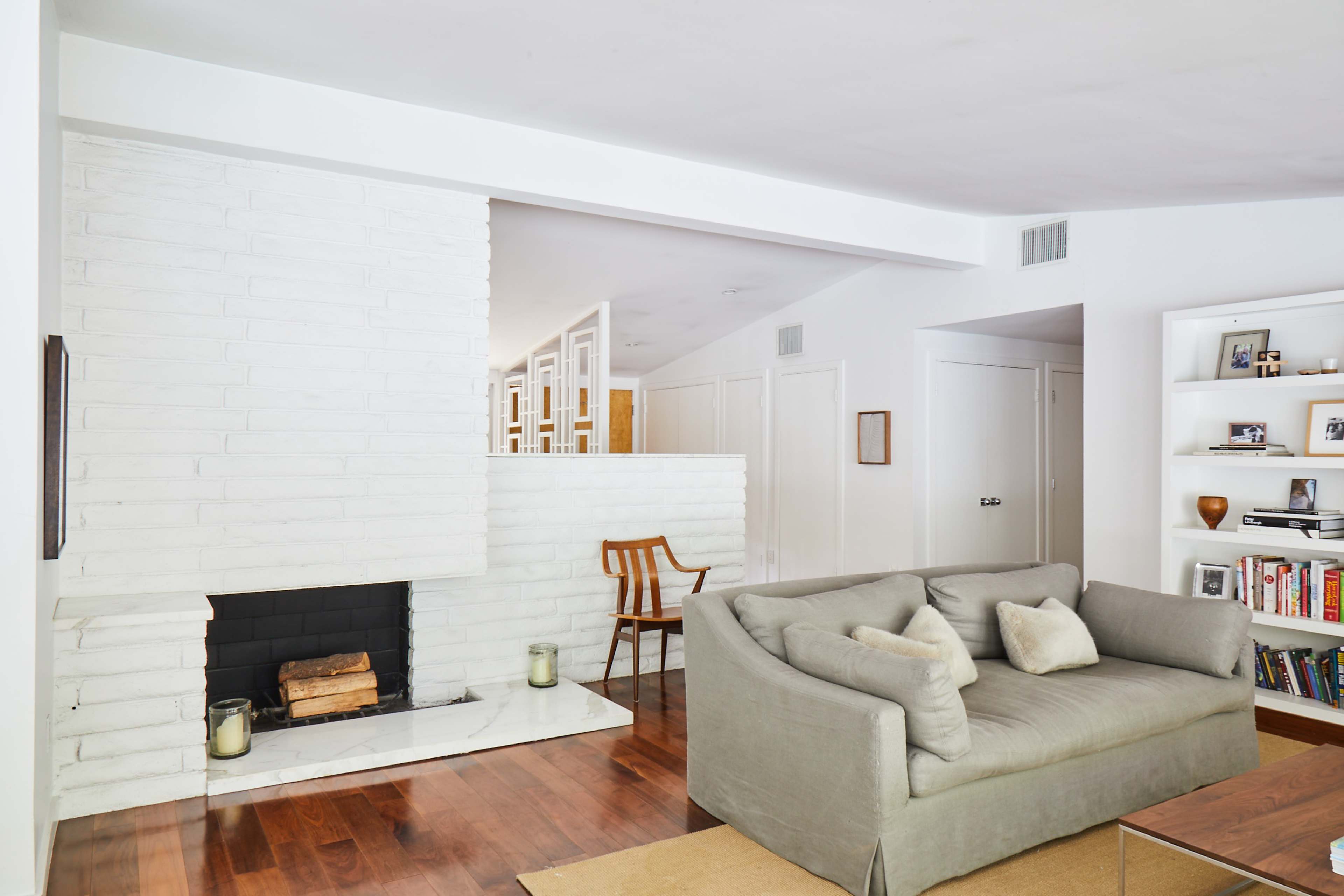 A modern living room featuring a gray sofa, a white brick fireplace, and wooden flooring, with shelves displaying books and photographs.